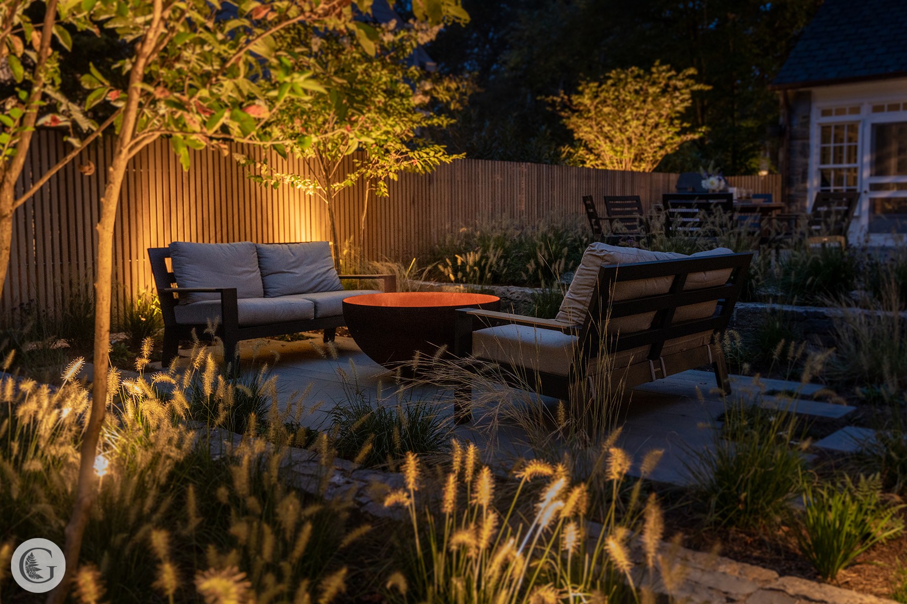Nighttime outdoor garden seating area with two benches, a table, and surrounding plants, illuminated by warm lighting.