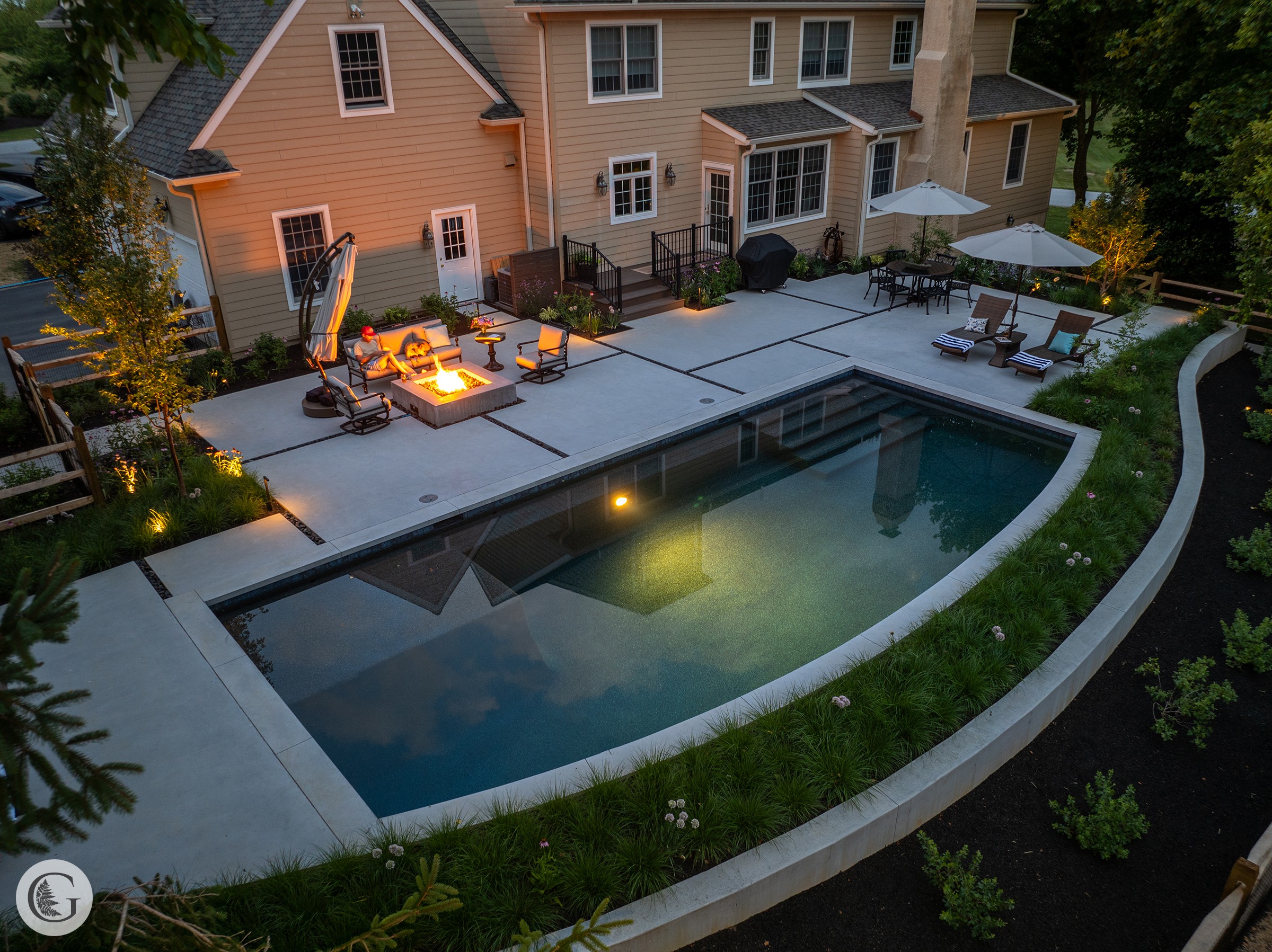 Backyard with swimming pool, patio area with fire pit, outdoor furniture, and a house in the background, illuminated at dusk.