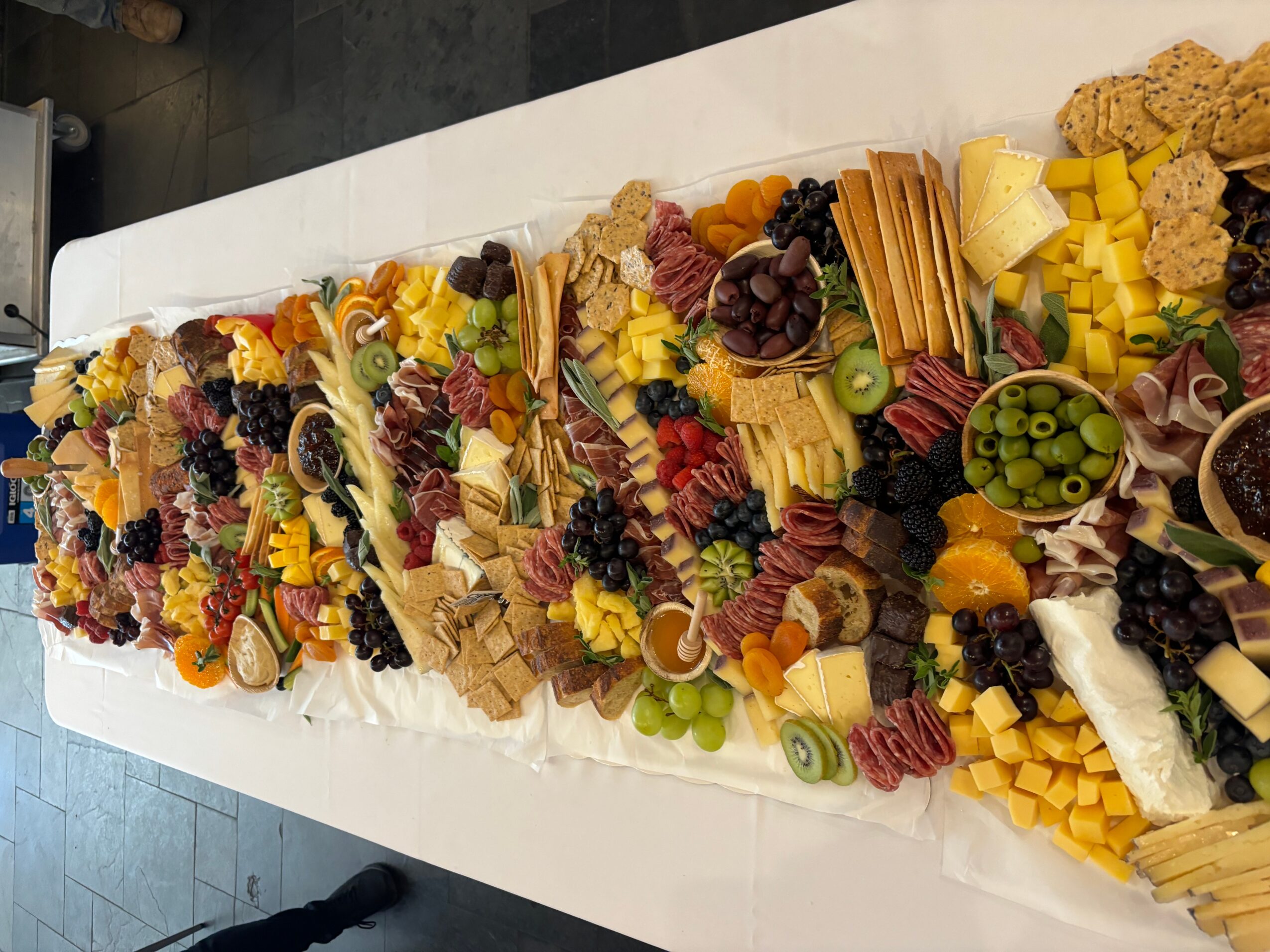 A long platter of assorted cheeses, fruits, nuts, and crackers arranged in sections on a white surface.