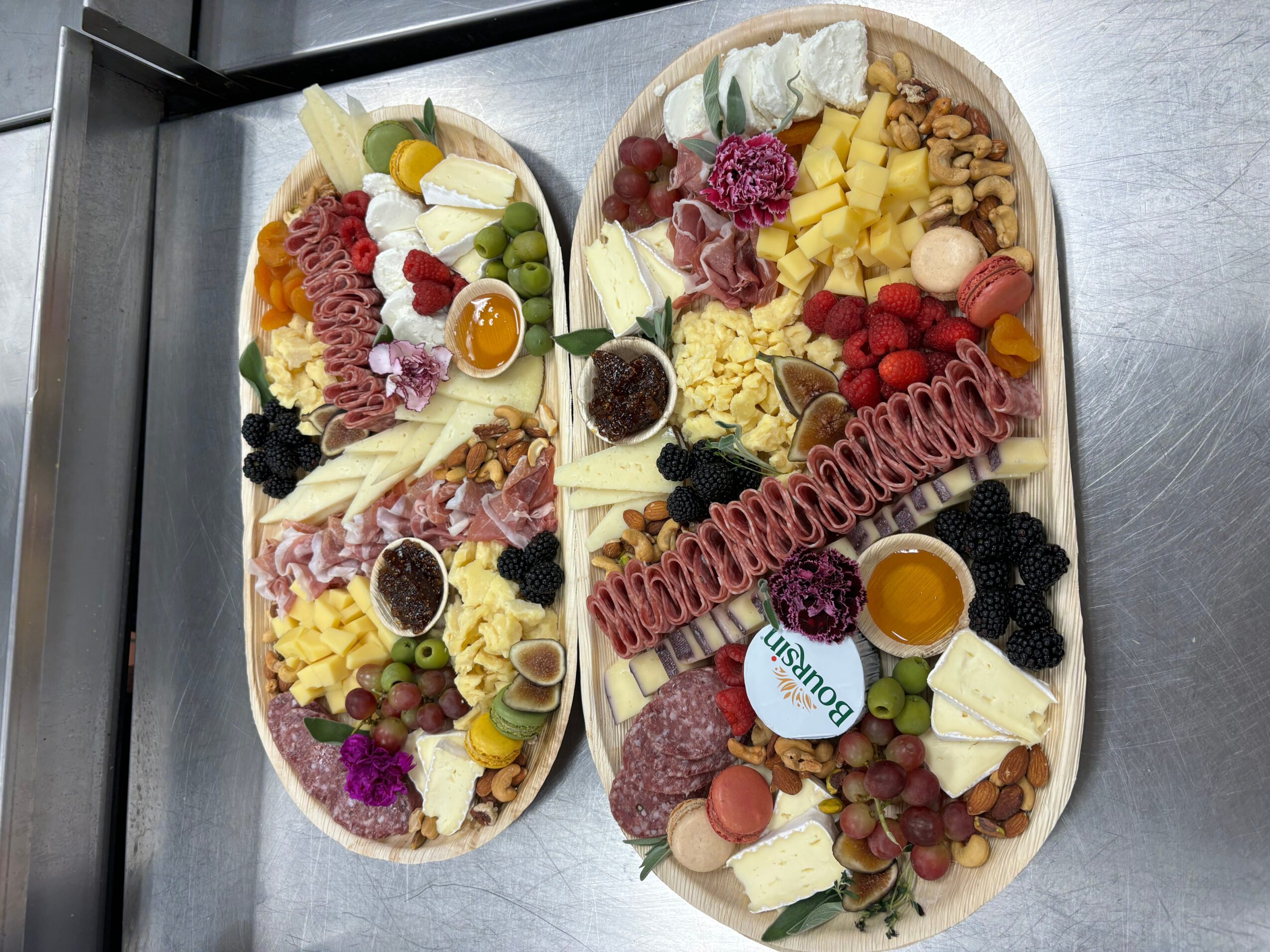 Two oval platters with assorted cheeses, fruits, and meats, garnished with herbs and edible flowers, on a stainless steel surface.