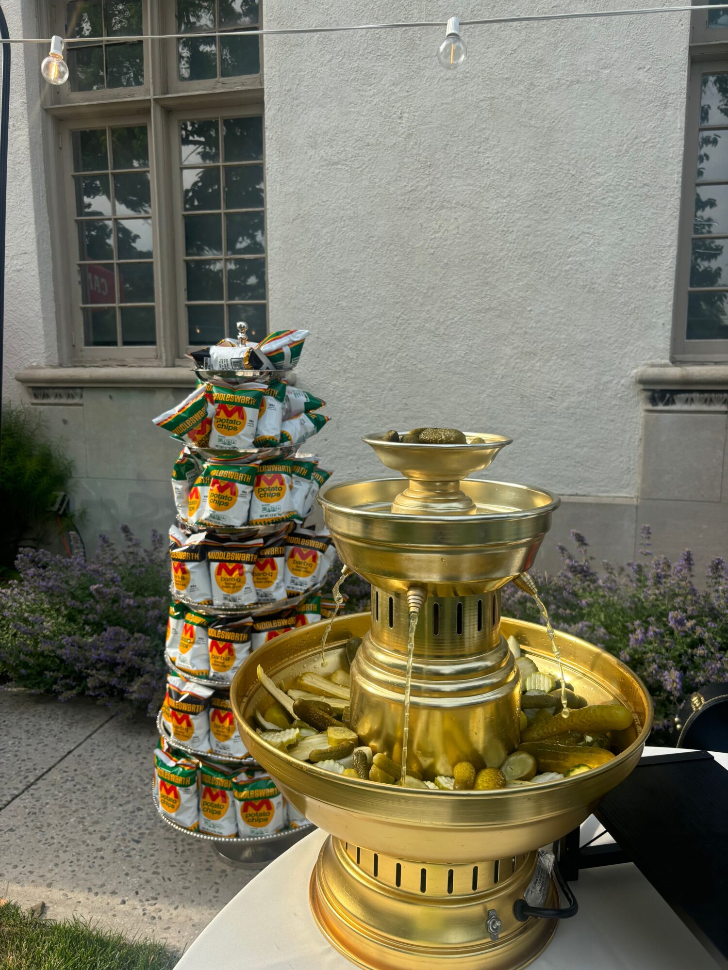 Golden decorative fountain with stacked colorful wrapped candies beside a building with large windows and purple flowers.