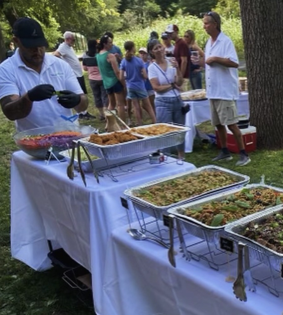 People gather outdoors around a table with food, serving dishes, and a person preparing food, in a park setting.