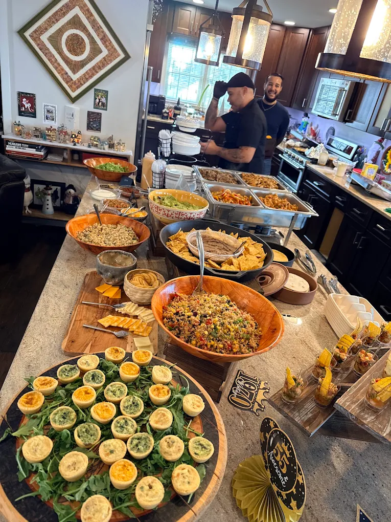 Buffet table with various dishes and a kitchen in the background, two people preparing food, decorated interior.