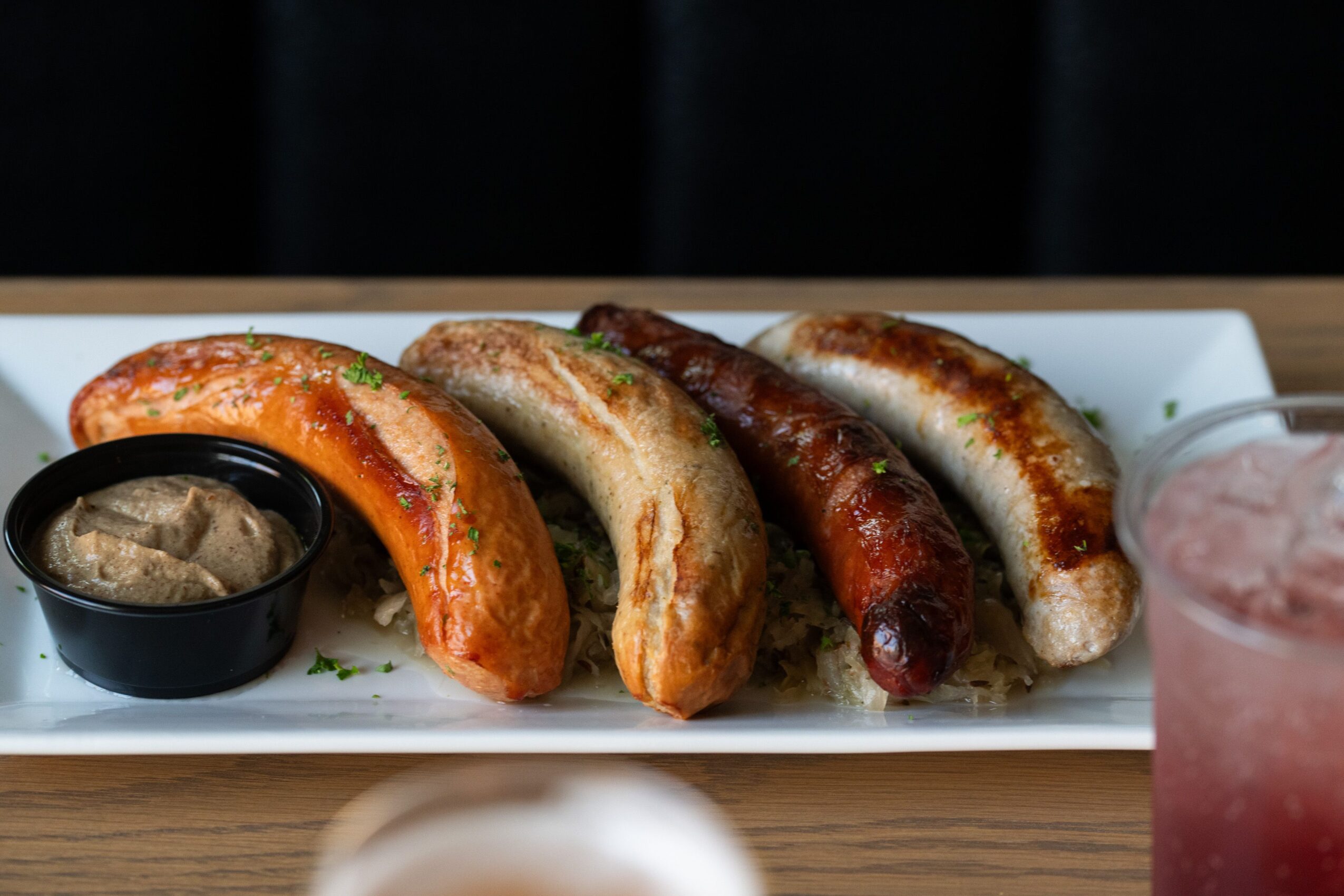 Five sausages on a white rectangular plate with a small black bowl of mustard, garnished with herbs, and a pink drink on the side.