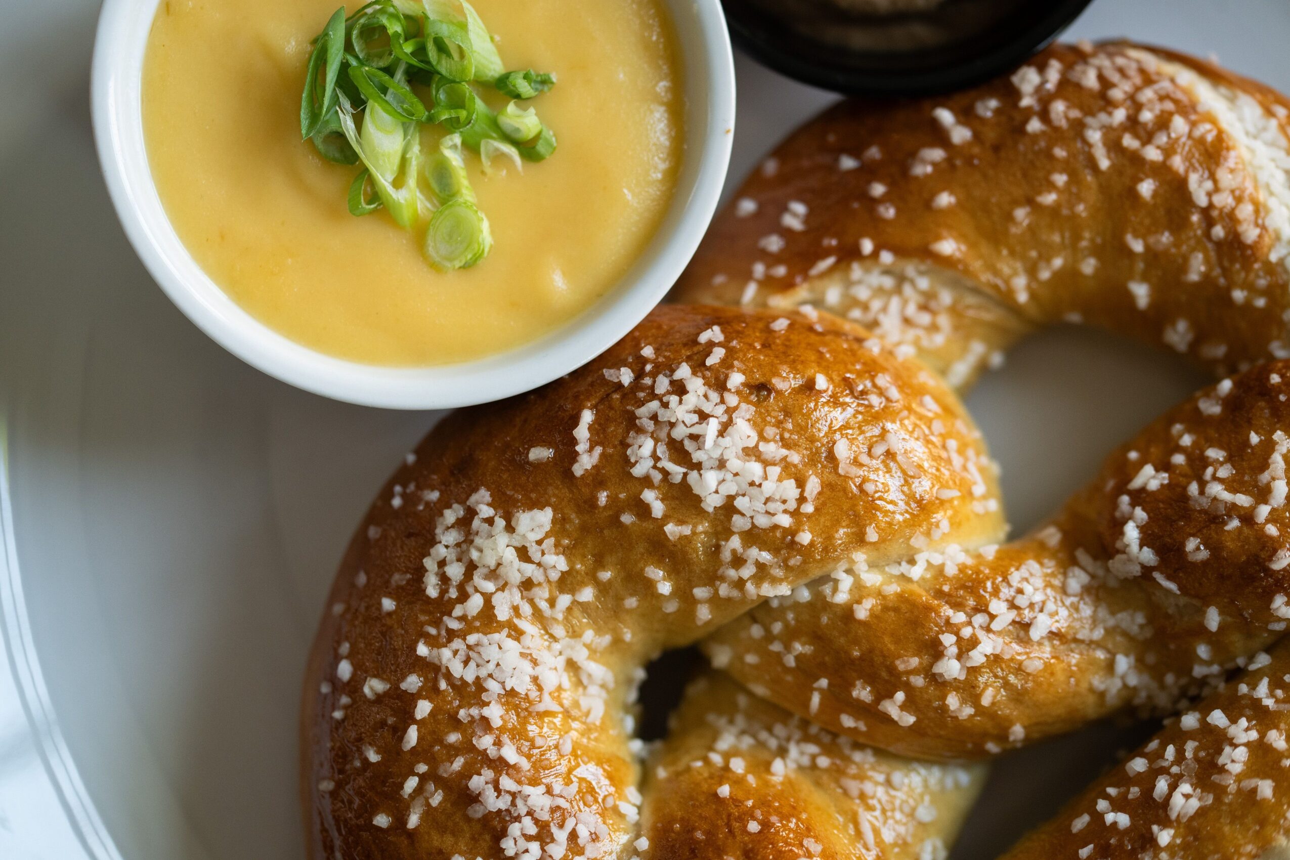 A pretzel with coarse salt and a small bowl of yellow mustard topped with chopped green onions.