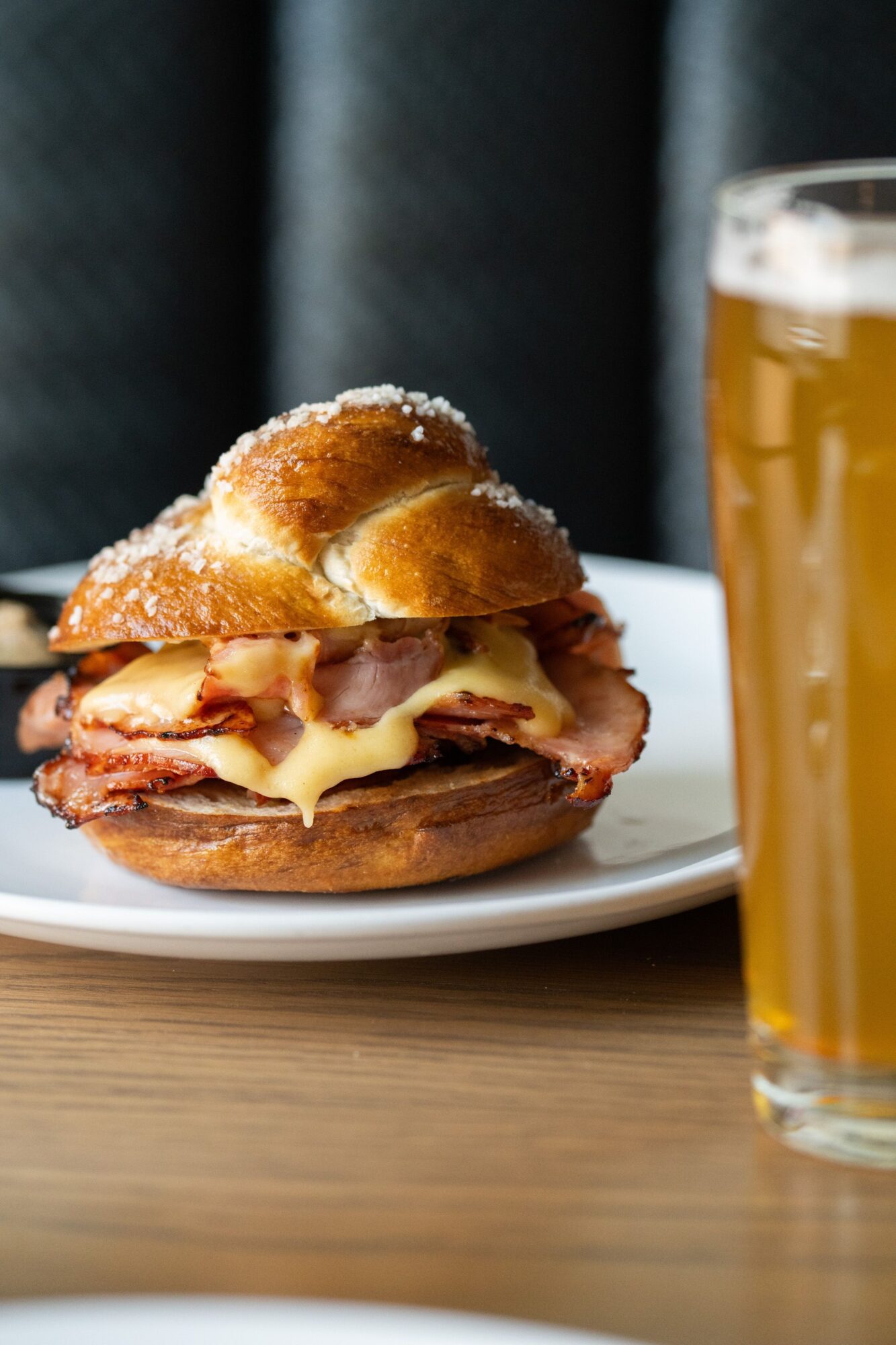 Sandwich with meat, cheese, and bread on a white plate, next to a glass of beer, on a wooden table.