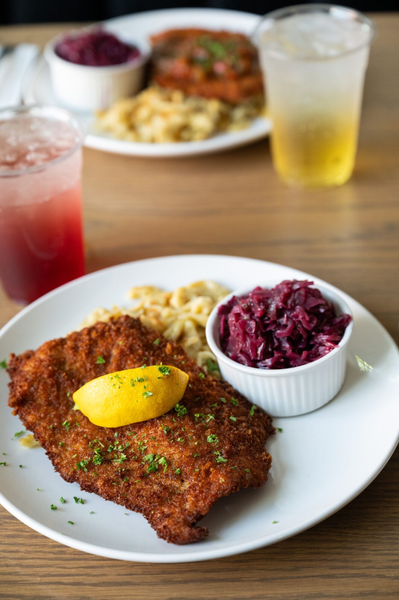 Fried fish fillet with lemon wedge, beet salad, and side of pasta on a white plate, with drinks in the background.