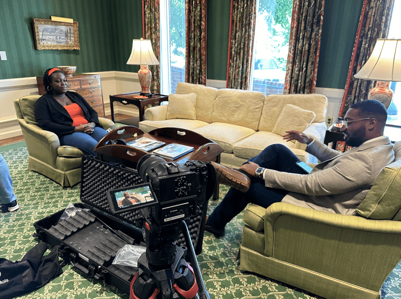 Two people sit on sofas in a room with green walls, curtains, and lamps, engaged in conversation, with camera equipment in foreground.