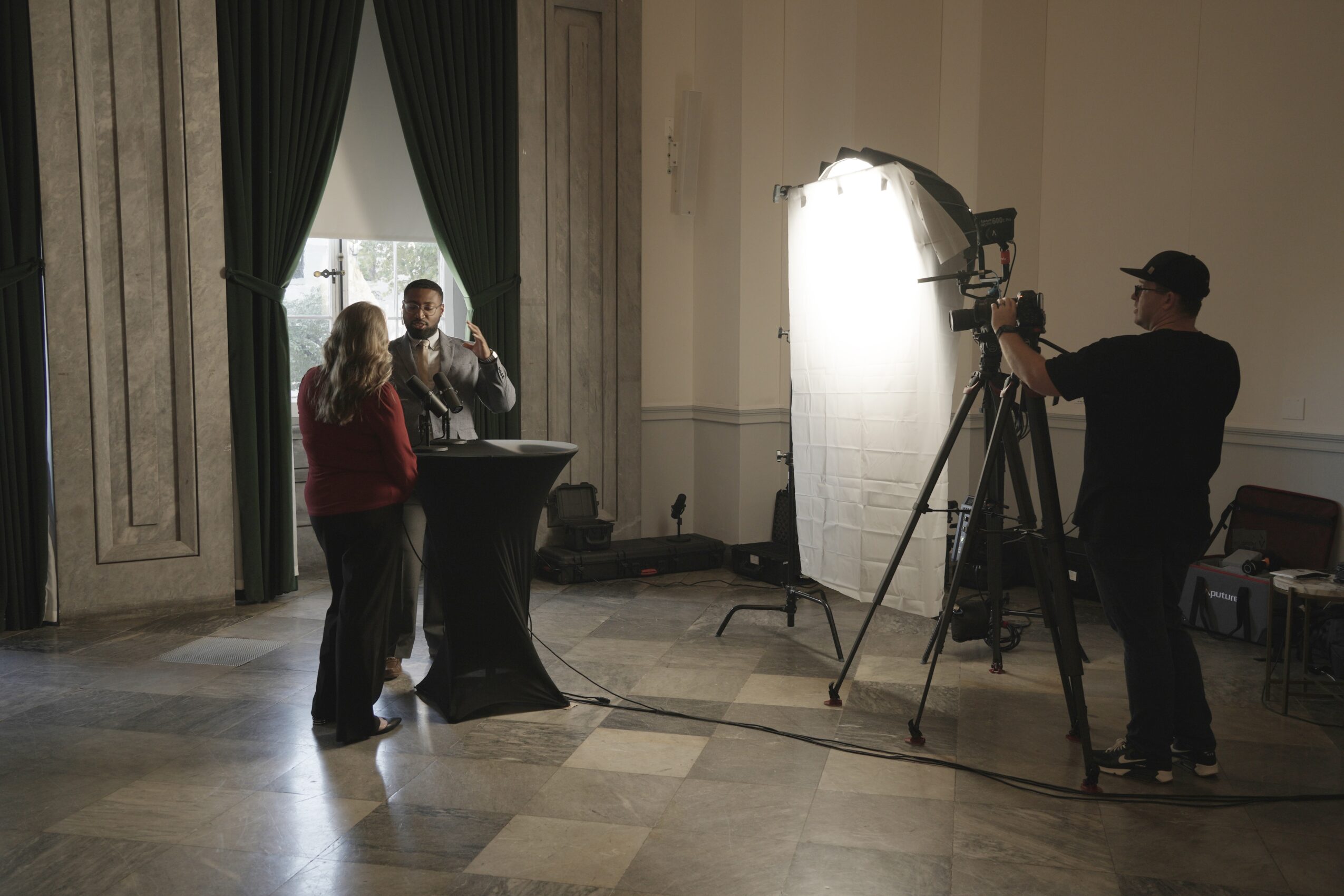 Two people are being filmed by a camera operator in a room with large windows and curtains, and a person standing nearby.