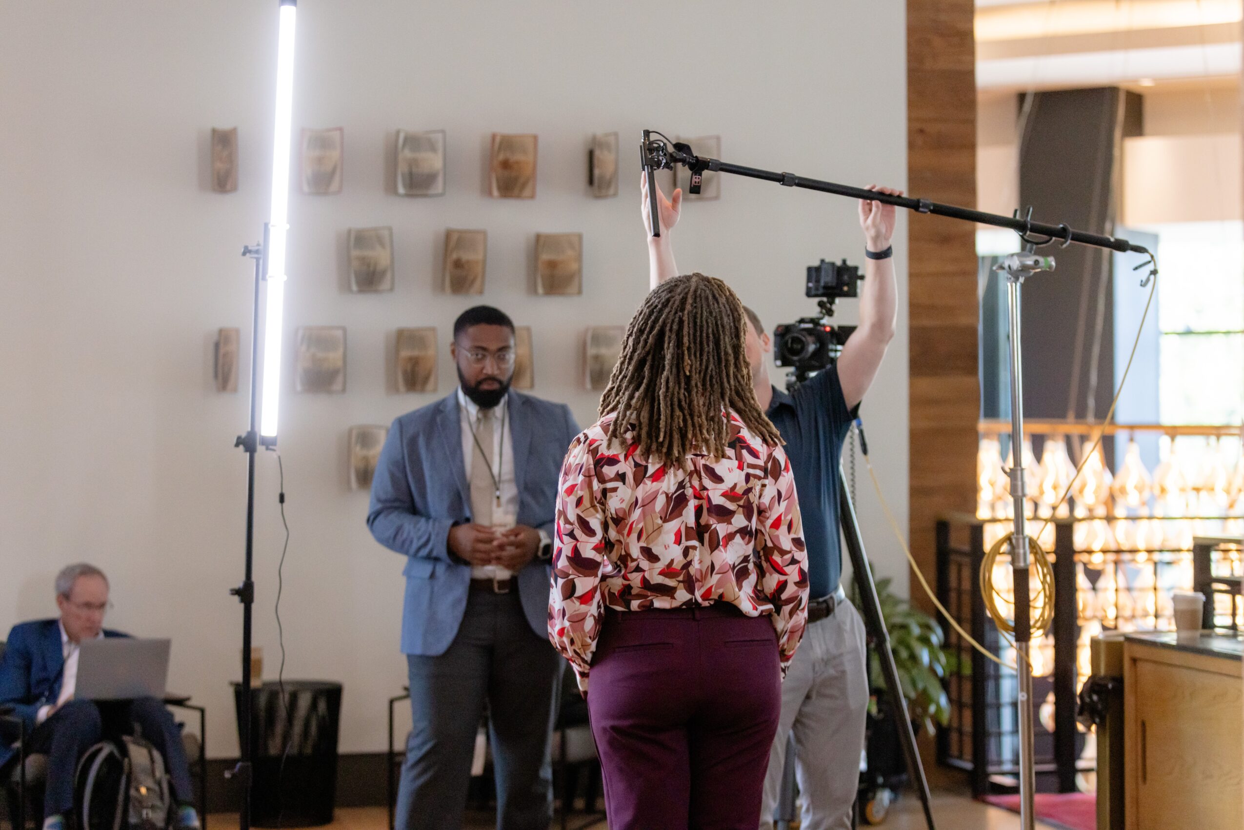 A woman with curly hair in a floral blouse and maroon pants is filming a man in a blue suit with a camera and crew in a bright indoor space.