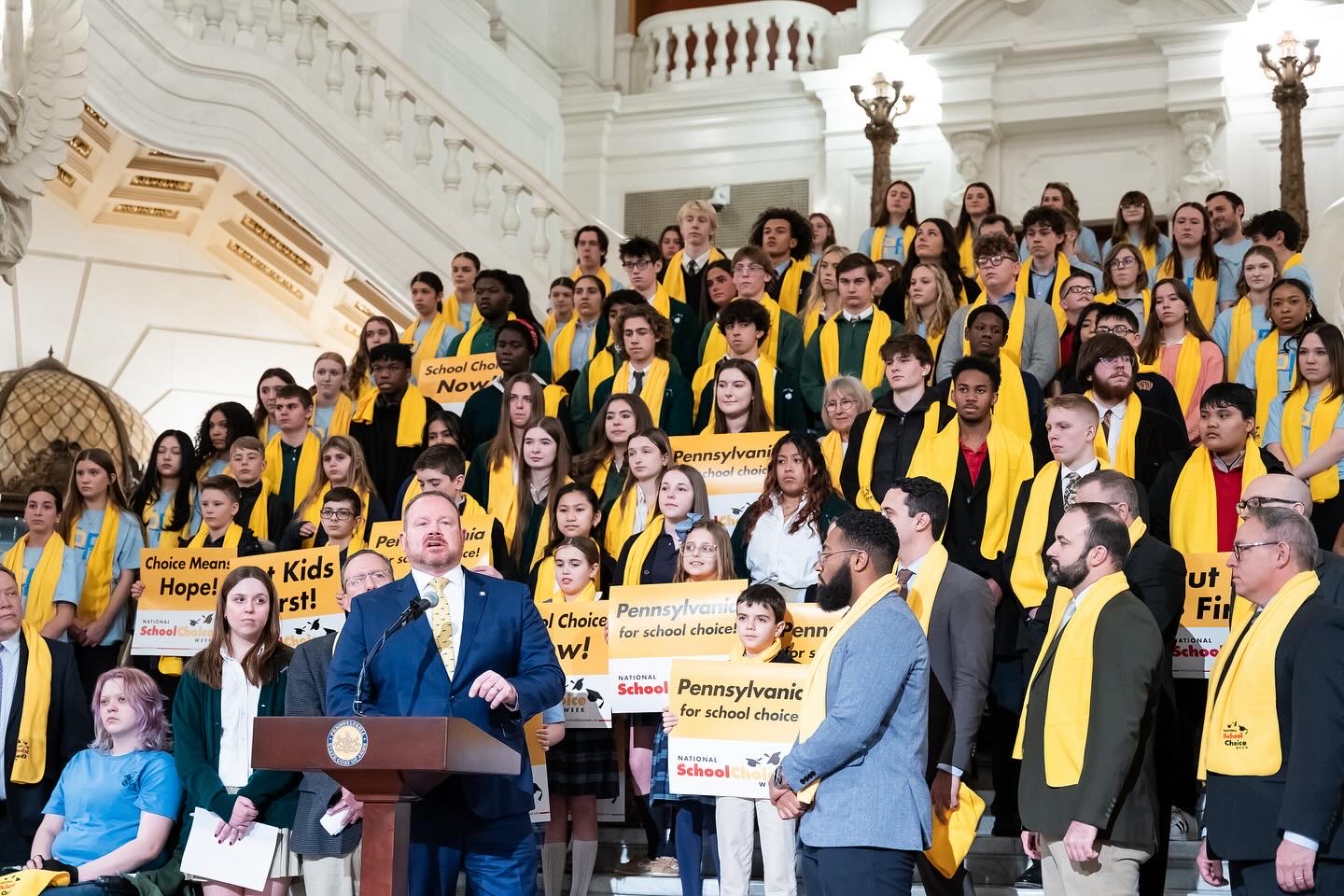 Group of students in yellow and black robes on stairs in a grand hall, holding signs and listening to a speaker at a podium.