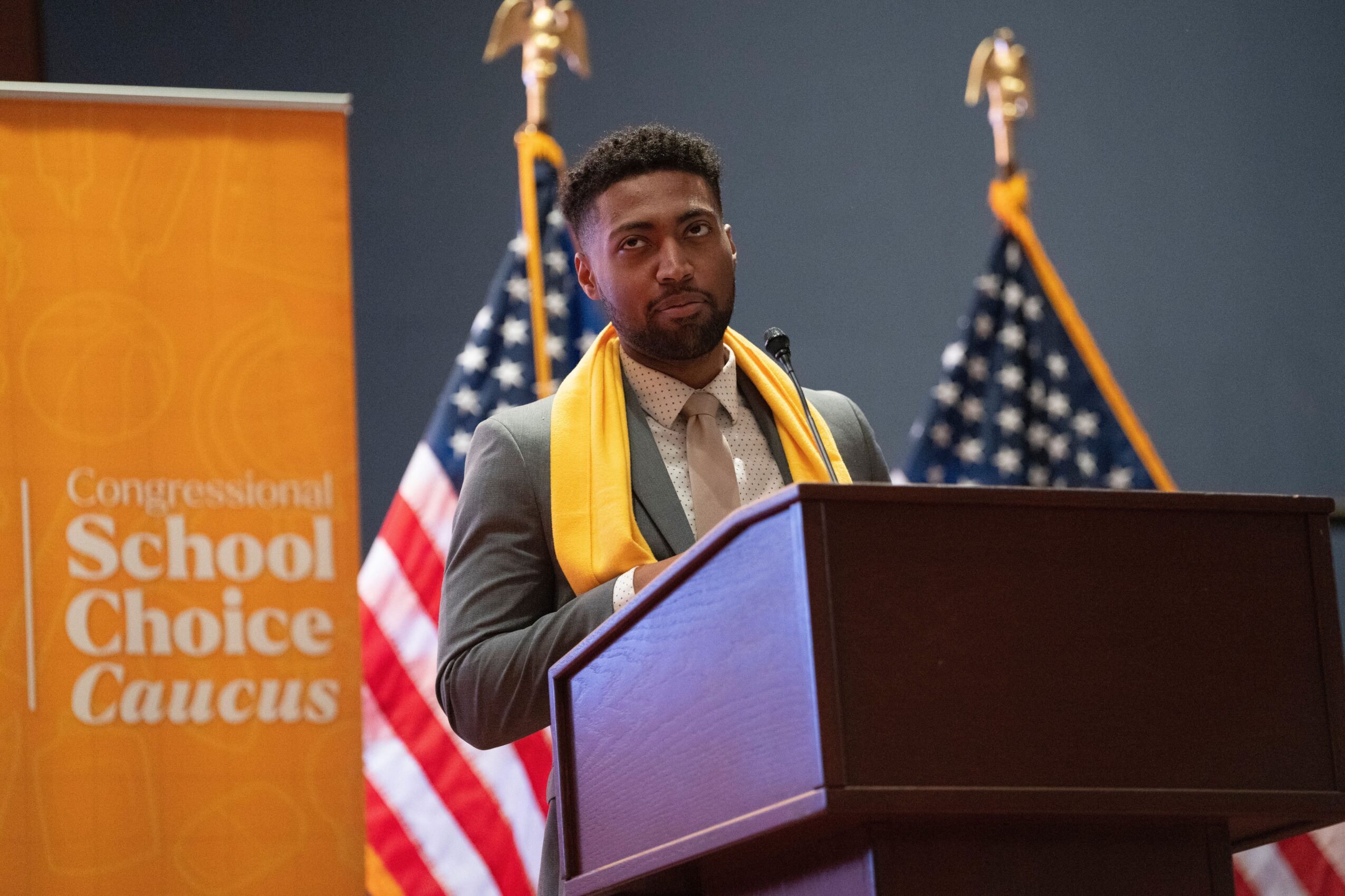 Man speaking at a podium with American flags behind him, orange banner reads 'Congressional School Choice Caucus'.