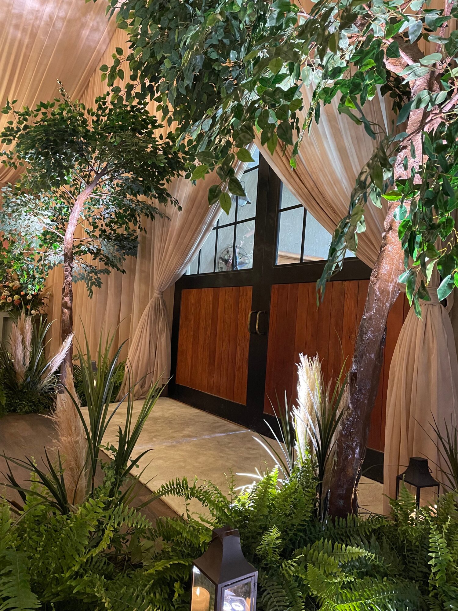 Indoor space with large trees, plants, and curtains, with a door in the background.