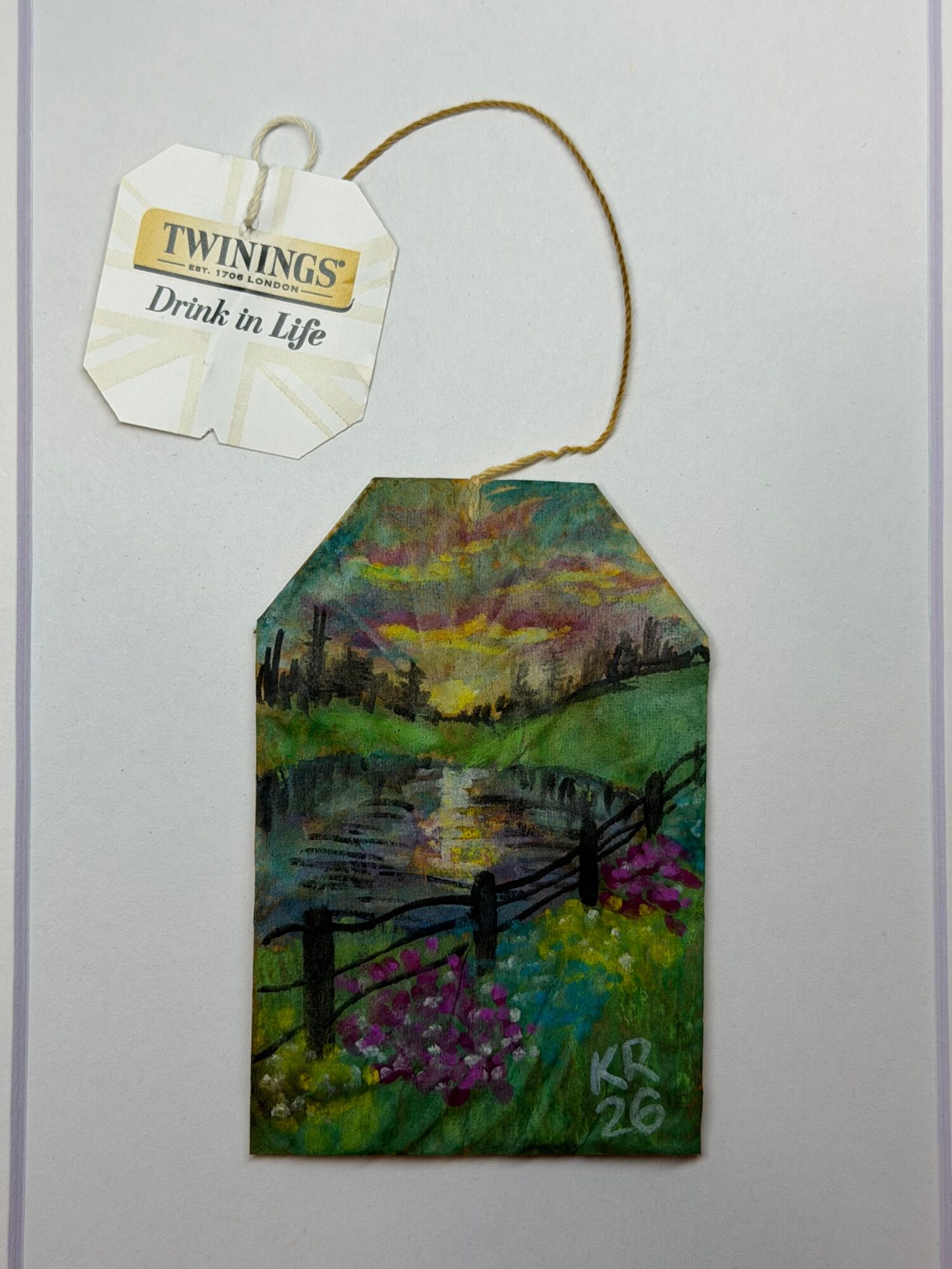 Colorful painted tag with landscape scene, flowers, and signature, attached to a string with a Twinings tea tag, on a white background.