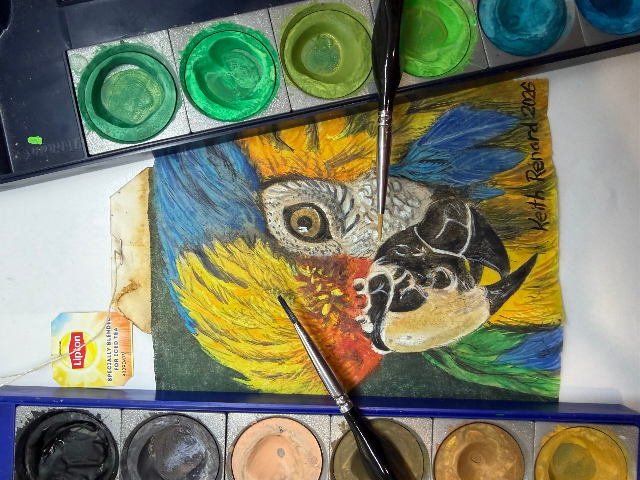 Colorful painting of a bird with yellow, blue, and black feathers, and a detailed eye, on paper with paint supplies nearby.