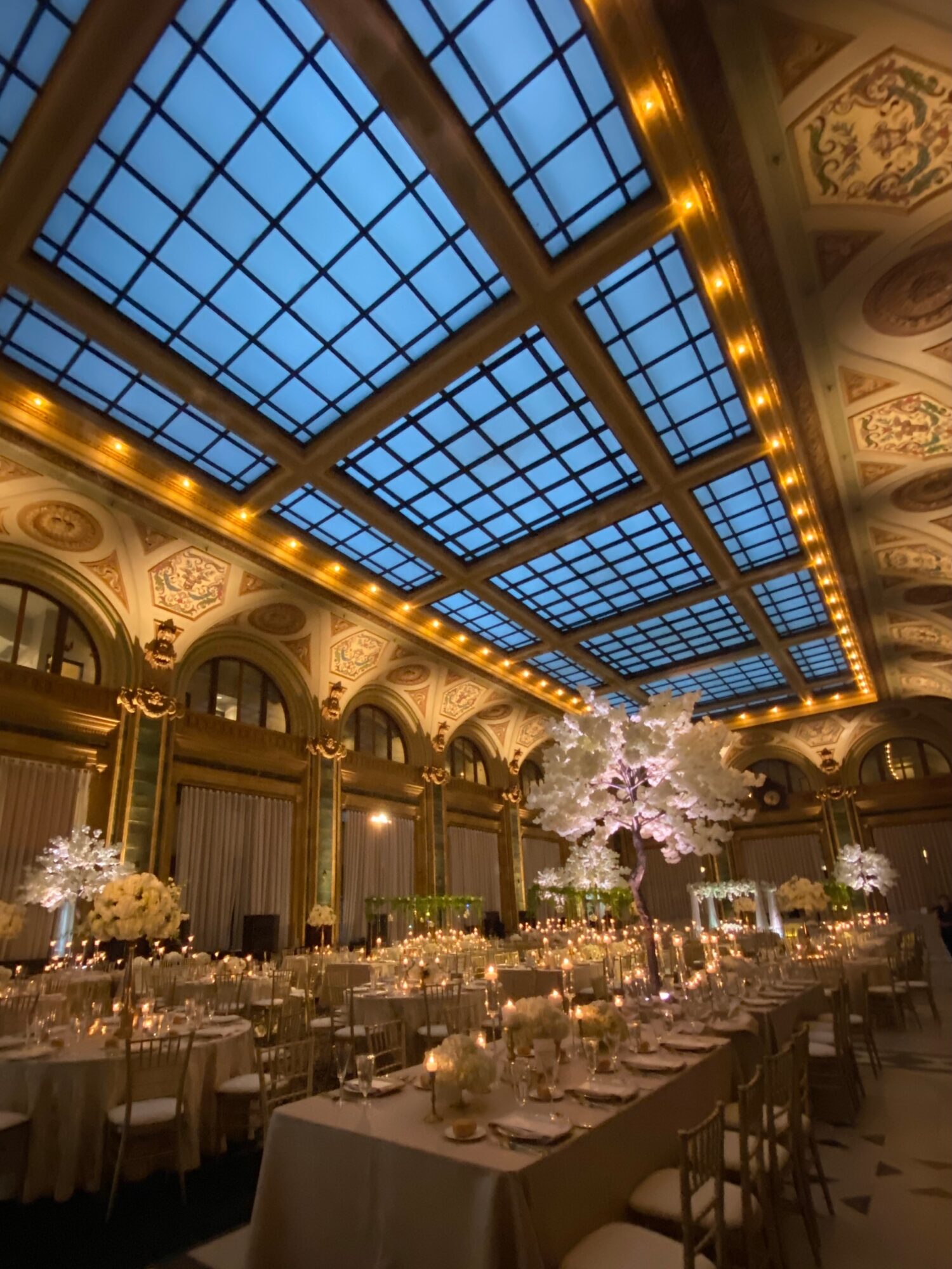 Elegant banquet hall with a large blue glass ceiling, ornate ceiling decorations, and decorated tables with floral centerpieces.