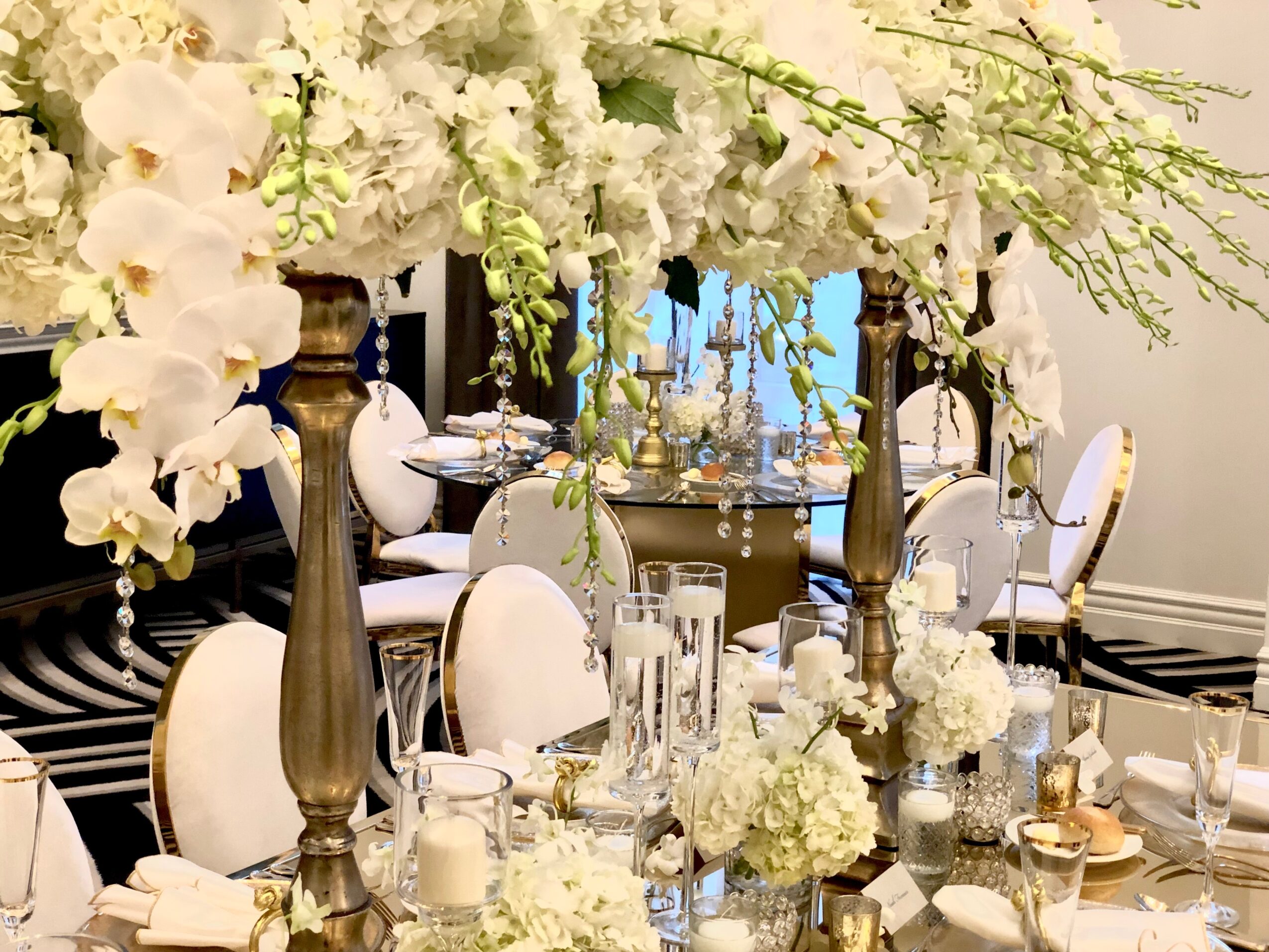 Elegant dining table decorated with white flowers, candles, and tall candlesticks, surrounded by white chairs with gold accents, in a bright room.