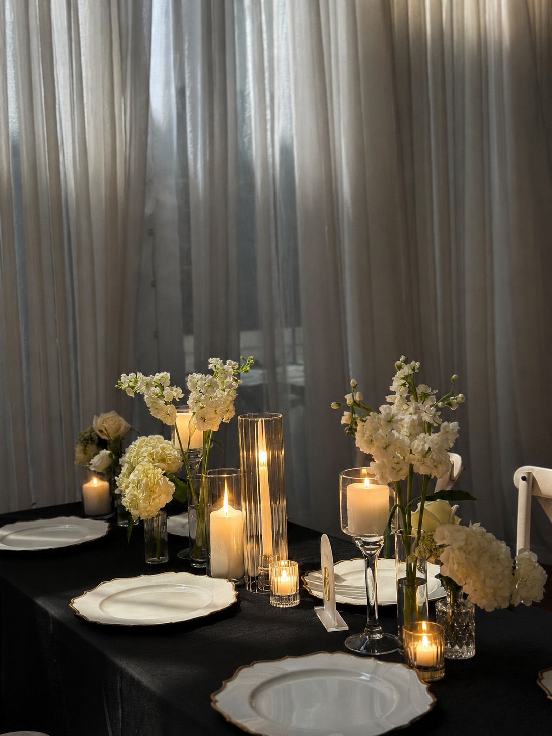 Table with floral arrangements, candles, and plates, set against curtains in a dimly lit room.