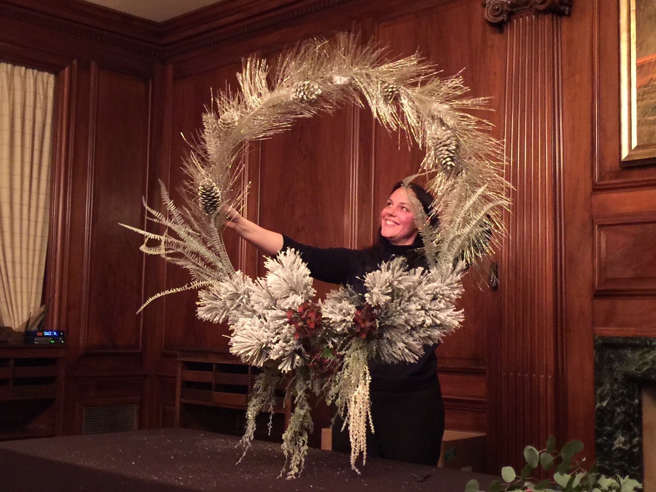 Woman holding a large decorative wreath made of dried plants and flowers indoors, smiling.