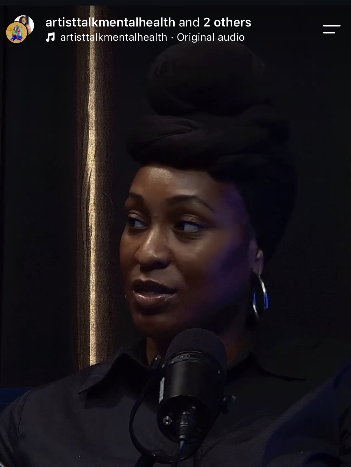 Woman with styled hair speaking into a microphone, dark background, wearing hoop earrings, looking to the side.