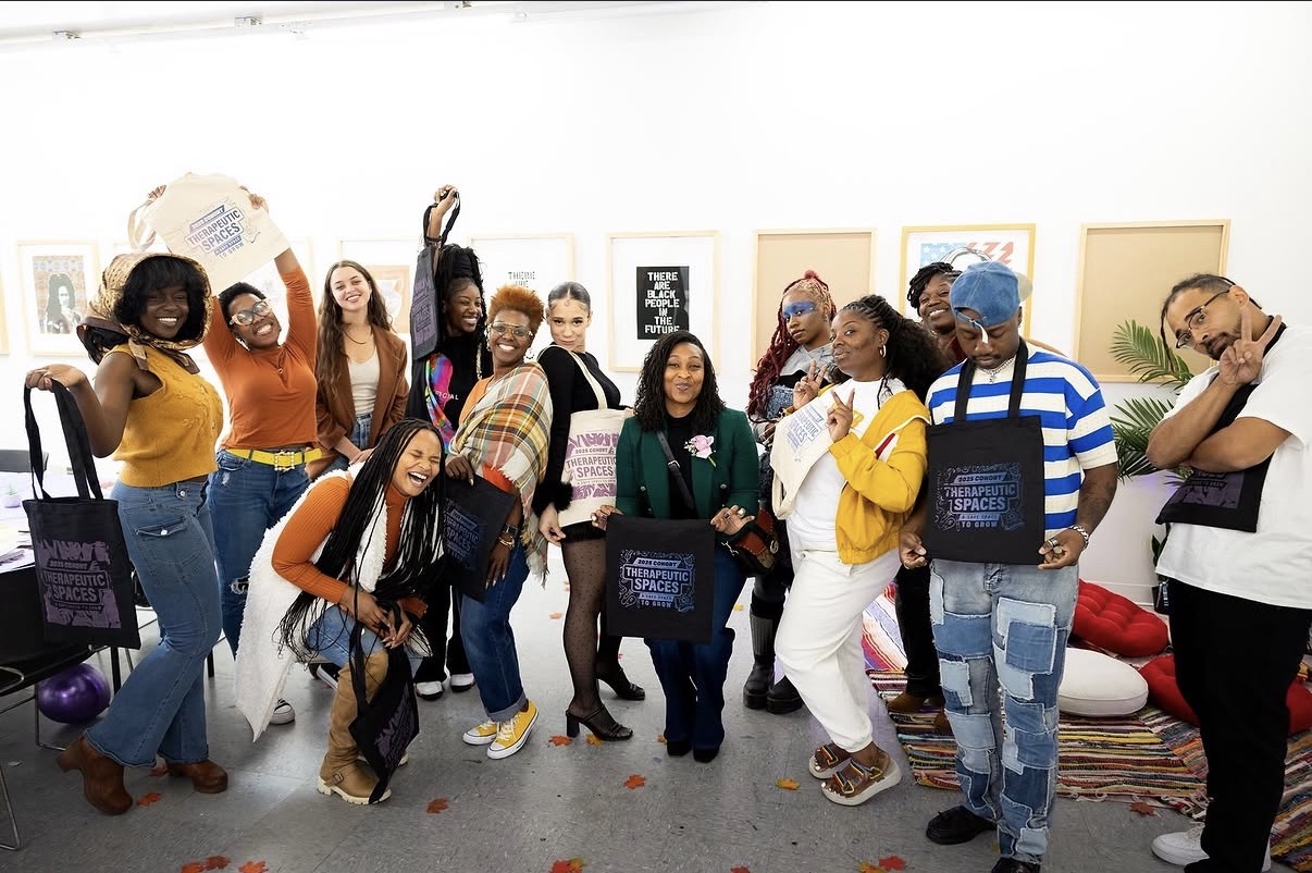 Group of diverse people posing in an art gallery, some holding bags, with framed artwork on the wall behind them.