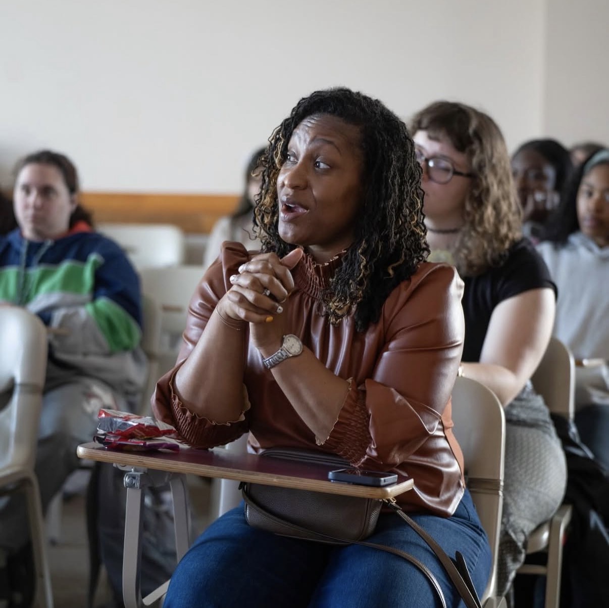 Woman with curly hair and brown leather jacket sitting in a classroom, listening attentively with hands clasped.