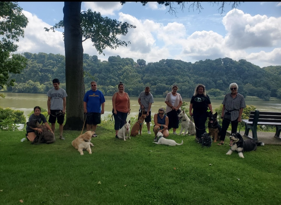 Group of people with dogs outdoors near a lake, trees, and hills, some standing, some sitting, on grass under a partly cloudy sky.