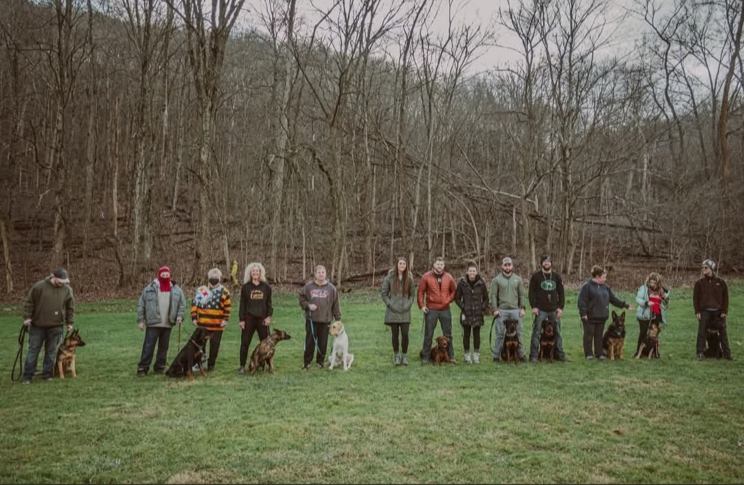 Group of people with dogs standing in a line on grass, trees in the background, overcast sky.