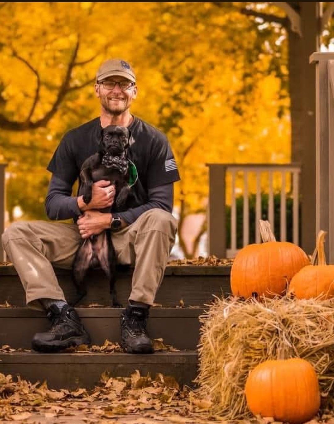 Man sitting on steps holding a puppy, surrounded by pumpkins and hay, with autumn trees in background.