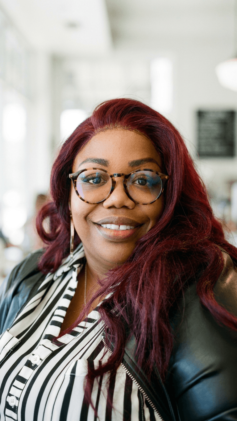 Woman with glasses and long, wavy red hair smiling indoors, background blurred.