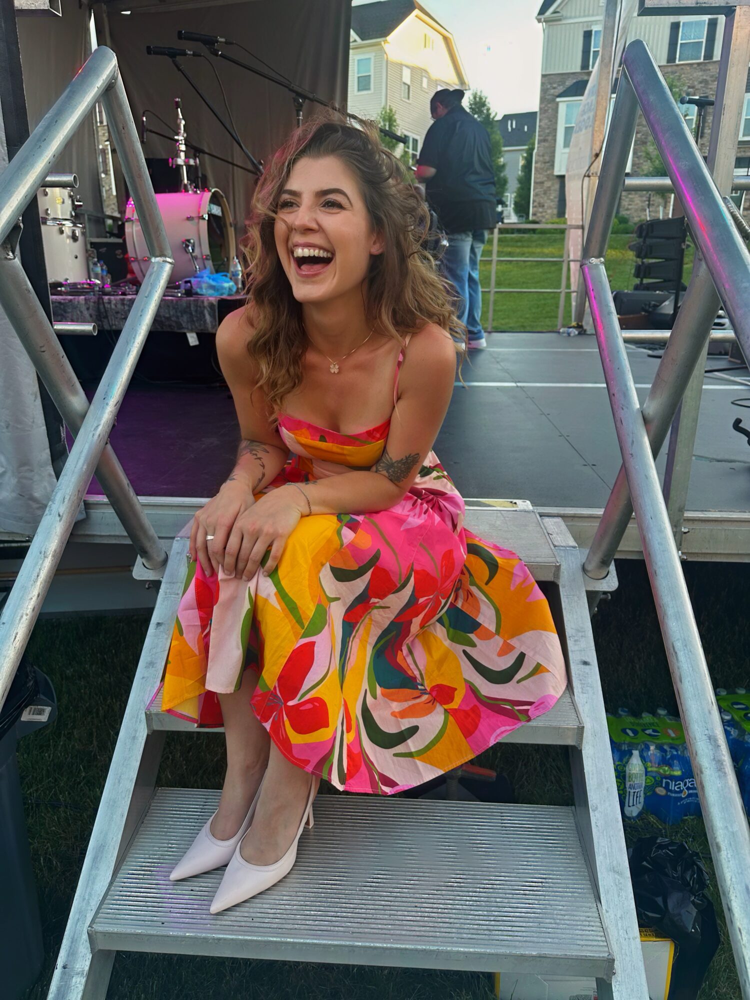 Young woman with curly hair laughing, sitting on stairs outdoors, wearing a colorful dress and white shoes.
