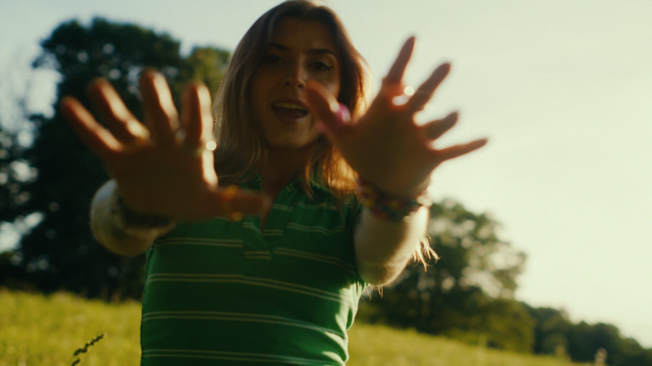 Person outdoors reaching towards the camera with hands, smiling, in a grassy area with trees and sky in background.