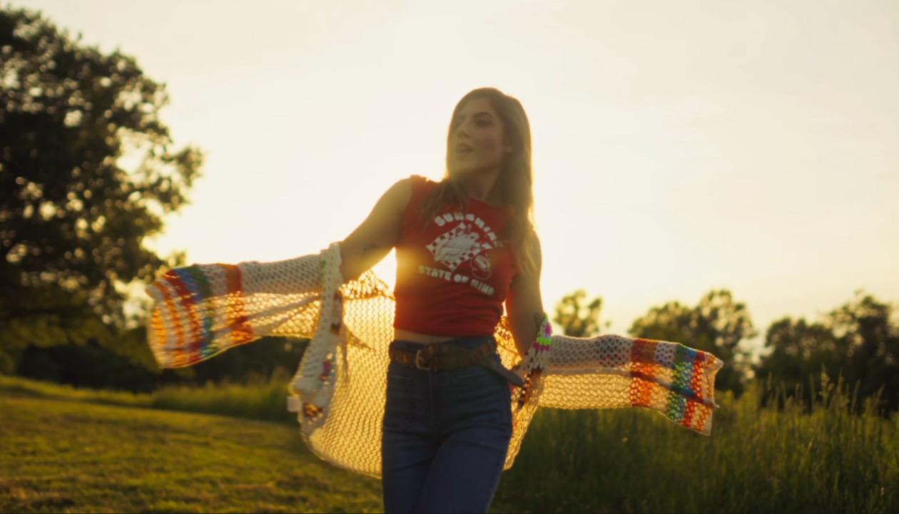 Person standing outdoors with arms outstretched, wearing a red top and jeans, in a grassy field with trees in background