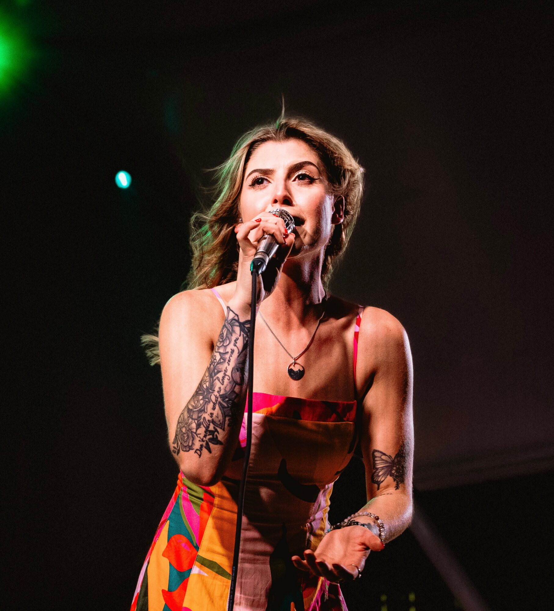 A woman with wavy hair sings into a microphone on stage, wearing a colorful dress and tattoos on her arms.
