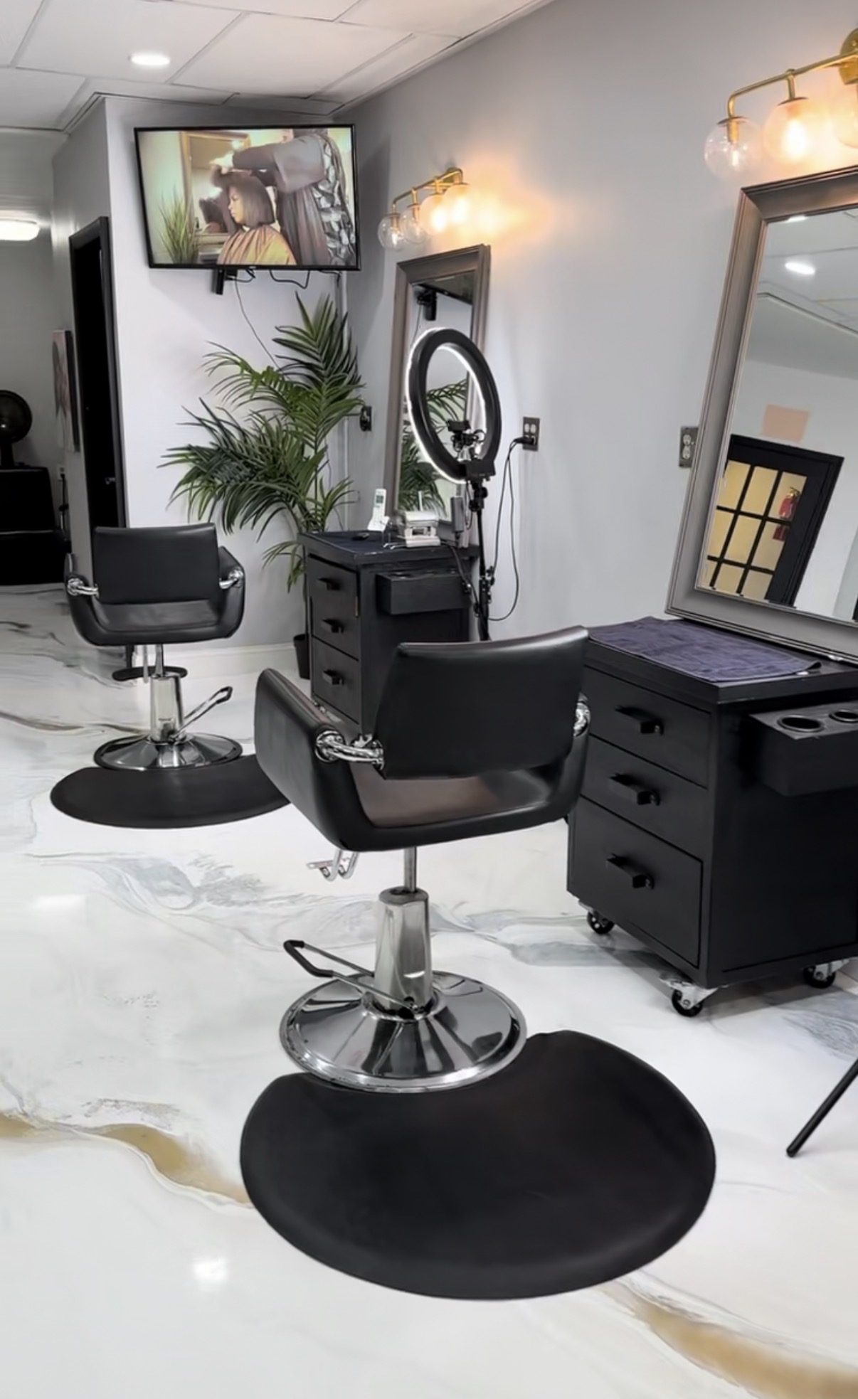 Salon interior with two black styling chairs, a mirror, a TV, and a potted plant, with white marble flooring.