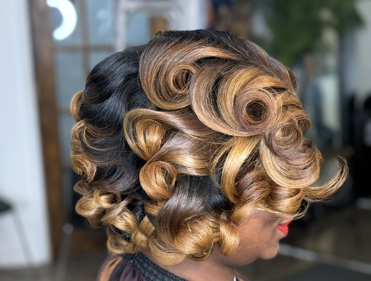 Side view of a woman with styled, voluminous curls in black and blonde hair, indoors with blurred background.
