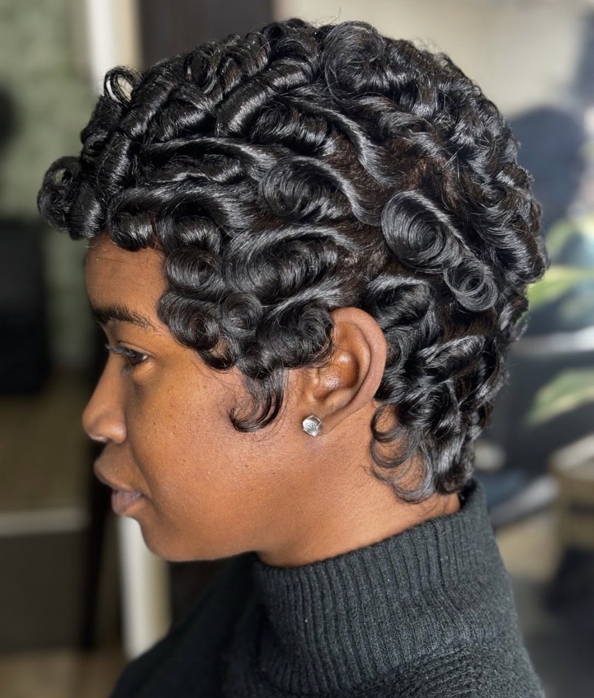 Side profile of a woman with short, curly, styled hair, wearing a black turtleneck and a small earring.