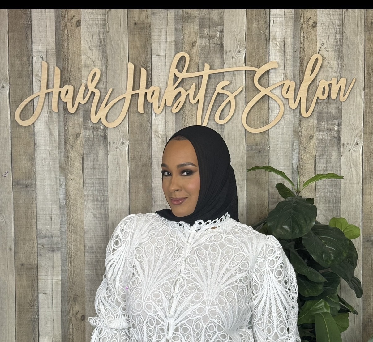 Woman wearing a white lace dress and black headscarf standing in front of a wooden wall with a sign that reads 'Hair Hubt's Salon' and a green plant.