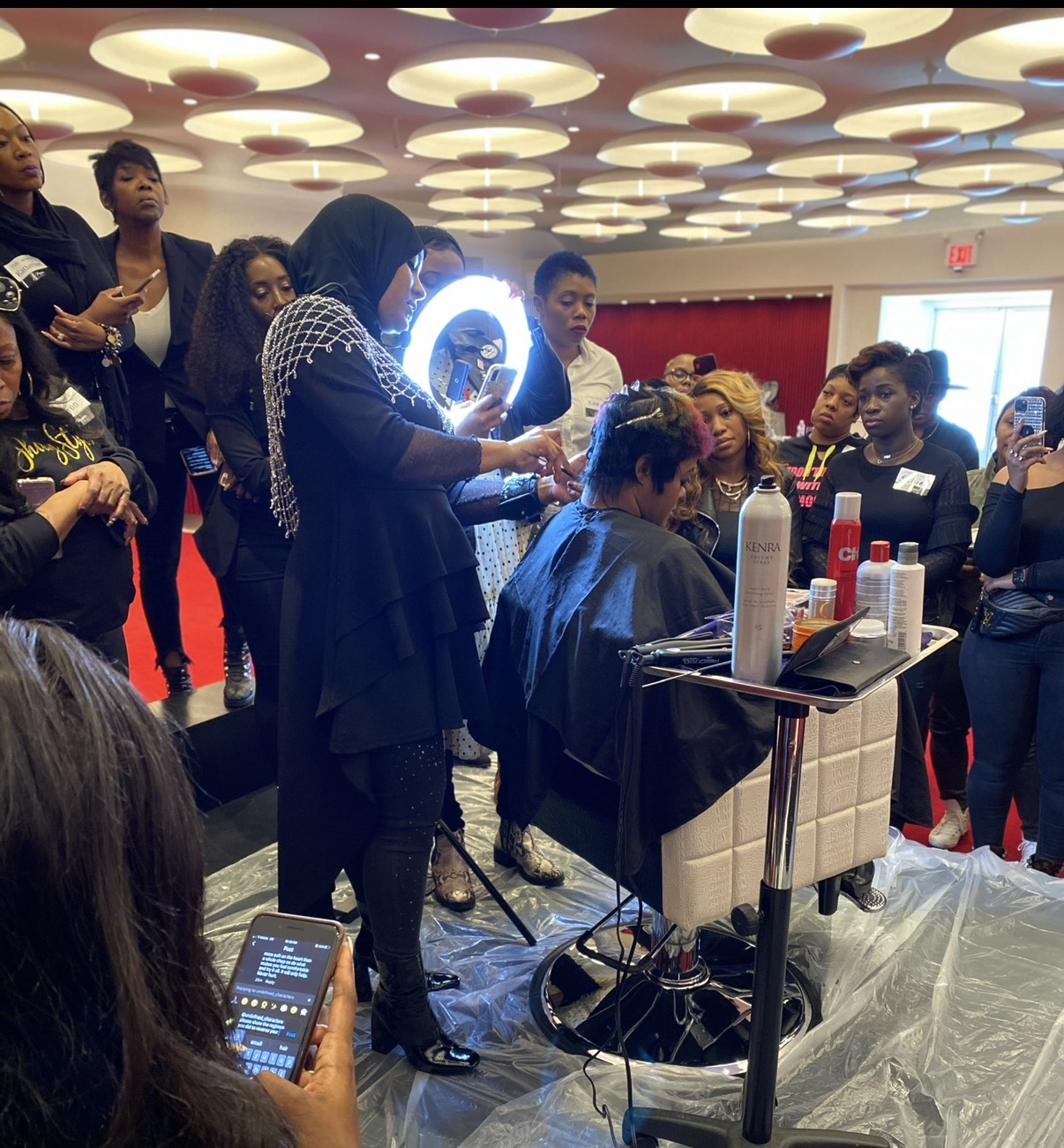 Group of people gathered around a person getting a haircut in a salon, with hair products on a trolley, ceiling lights overhead.