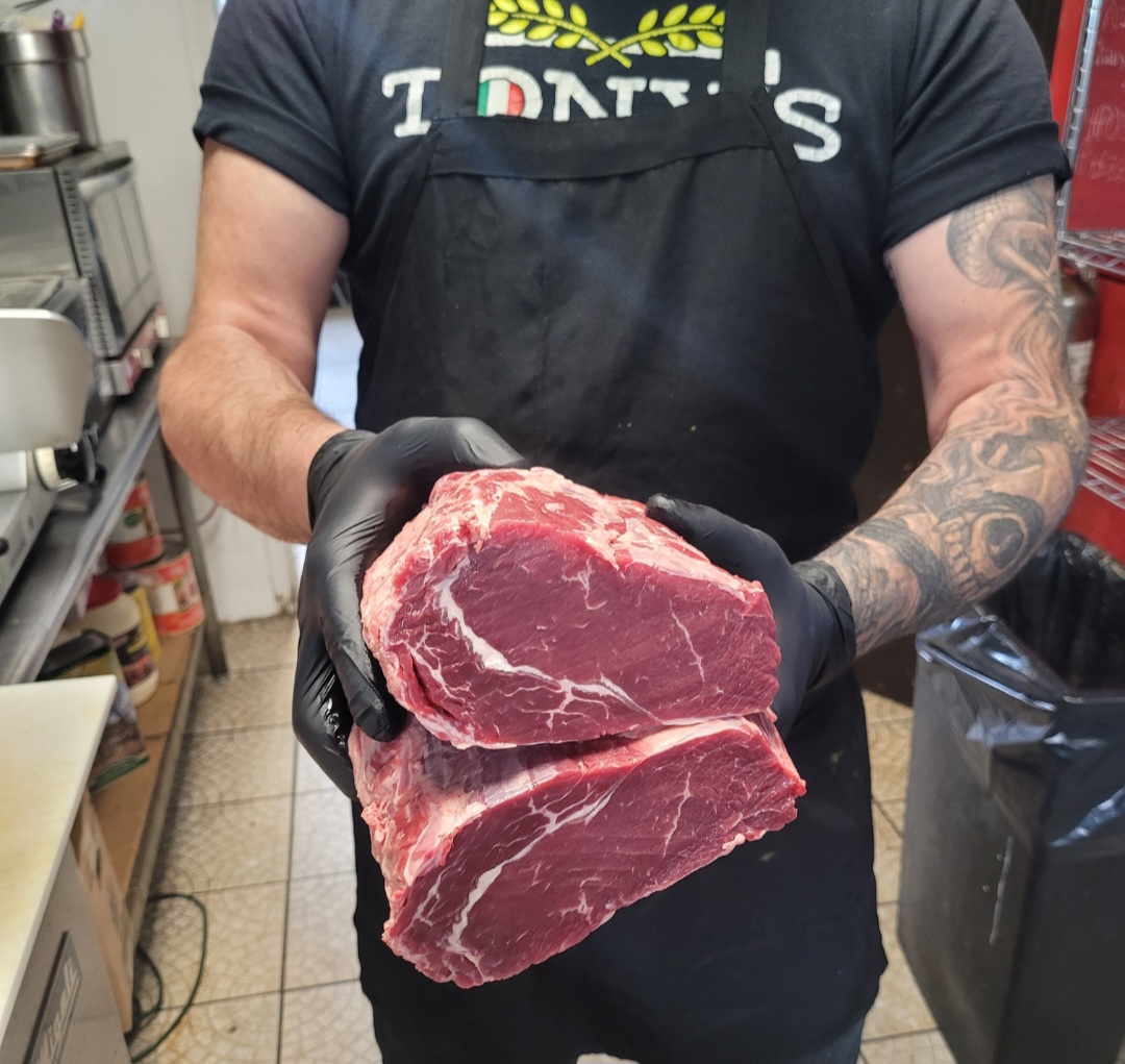 Person holding two large cuts of raw beef in a kitchen, wearing black gloves and an apron, with tattoos on arms.