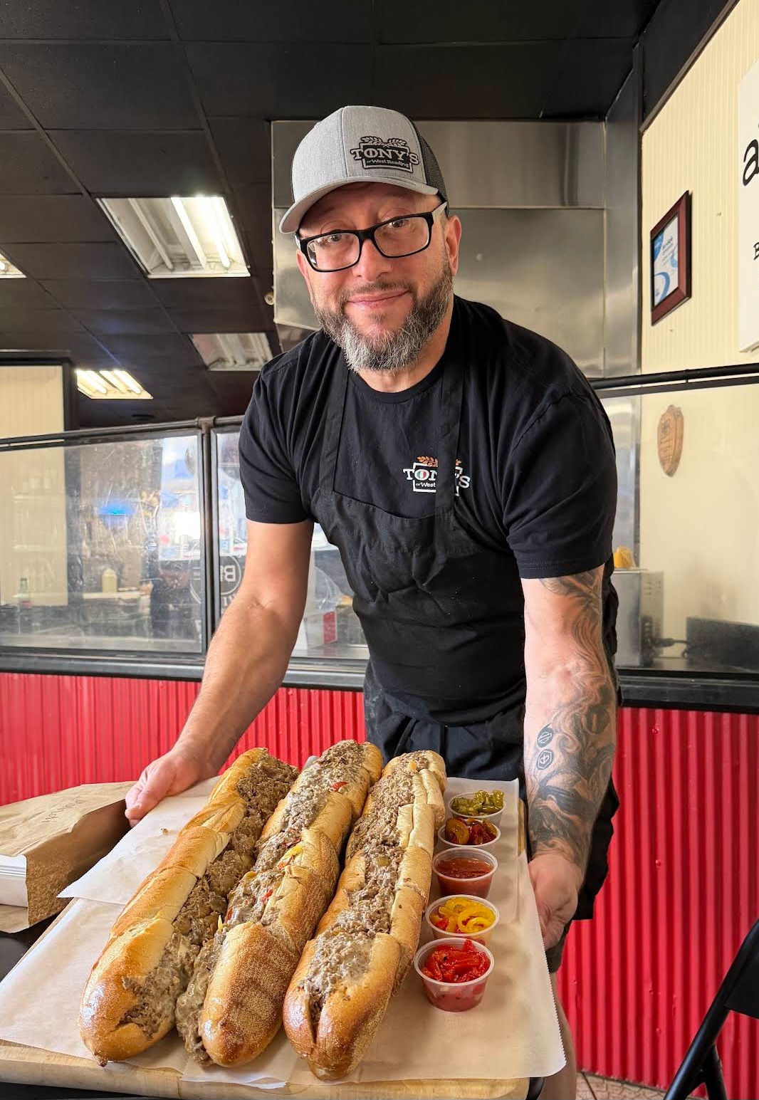 Man with glasses and a cap stands behind a tray of four large hot dogs with condiments, inside a restaurant.