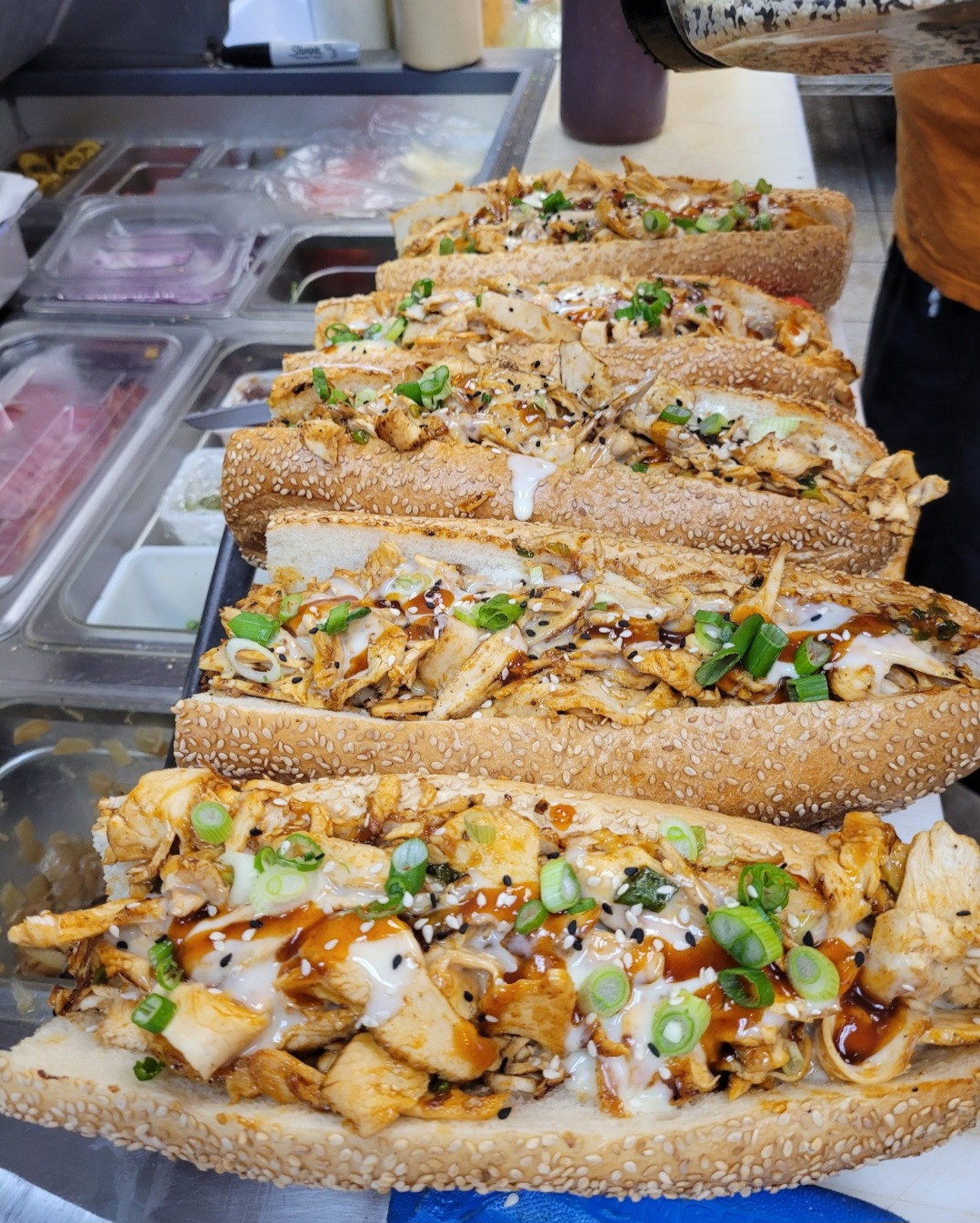 Six sandwiches with grilled chicken, green onions, and sauce on sesame seed buns, arranged on a counter.