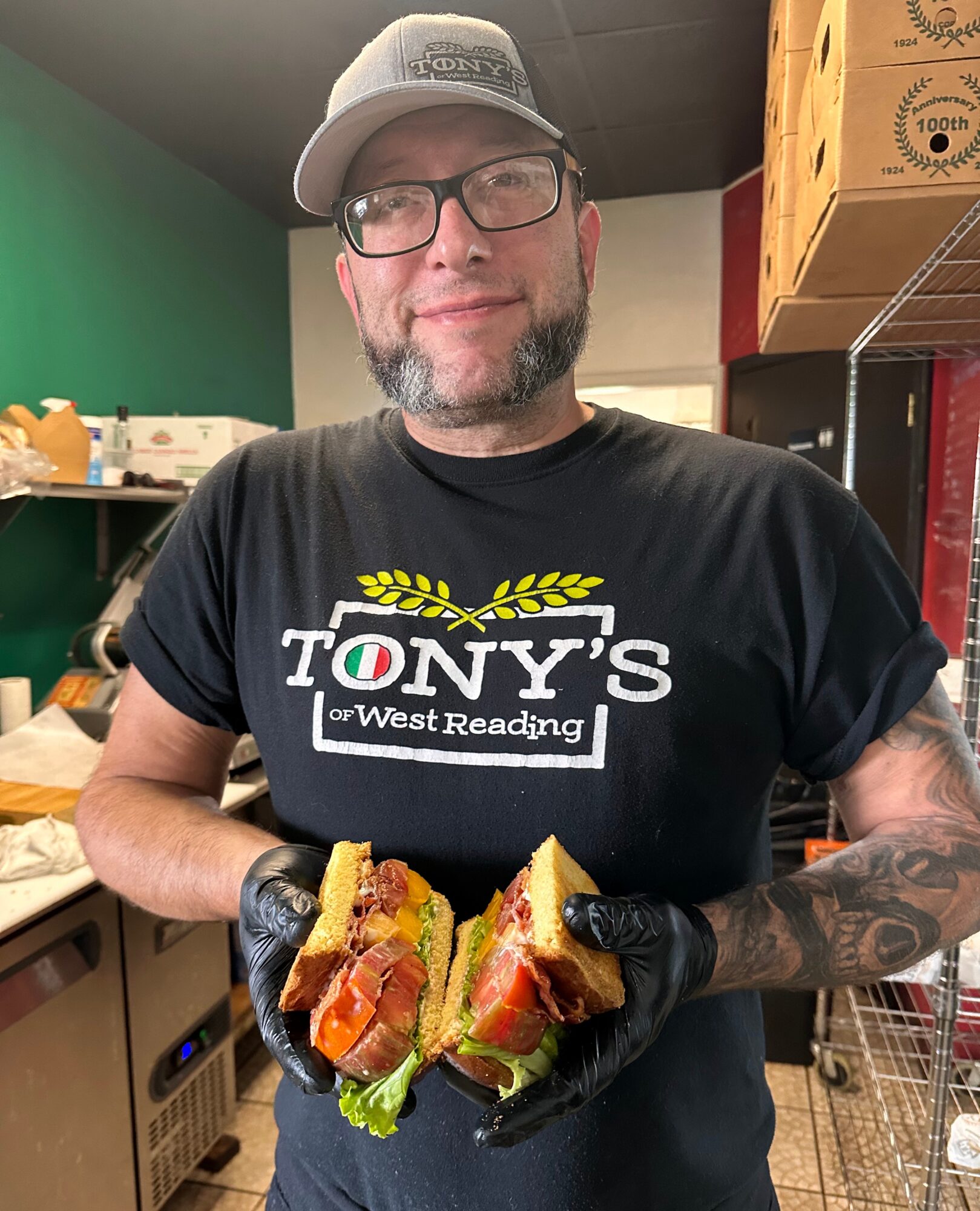 Man with glasses and a cap holding a sandwich in a kitchen, smiling, wearing a T-shirt with text, black gloves, shelves in background.