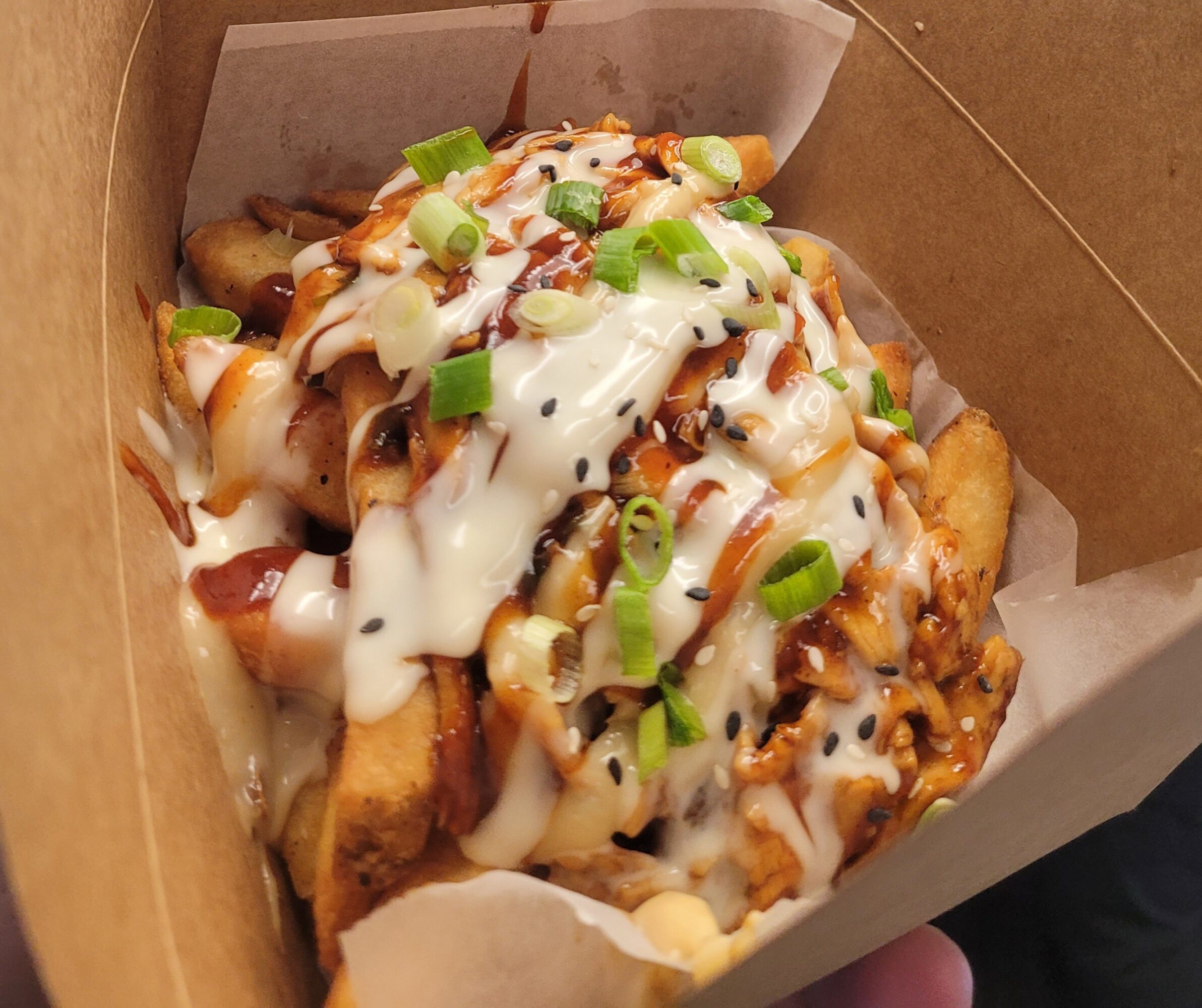 Fried chicken pieces topped with white sauce, chopped green onions, and drizzled with dark sauce inside a cardboard container.