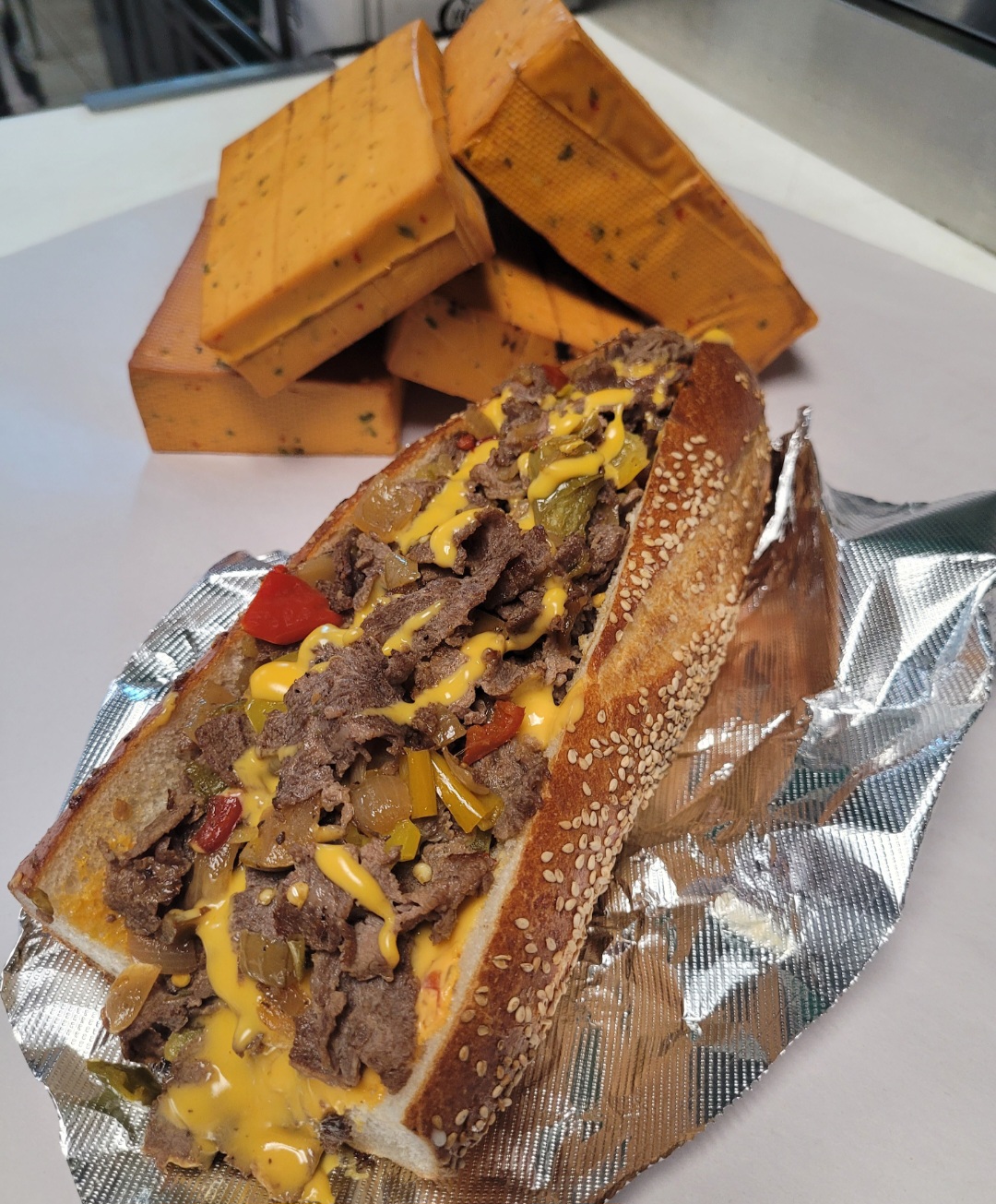 Sandwich with sliced beef, cheese, and toppings in a bun on foil, with cheese blocks in background.