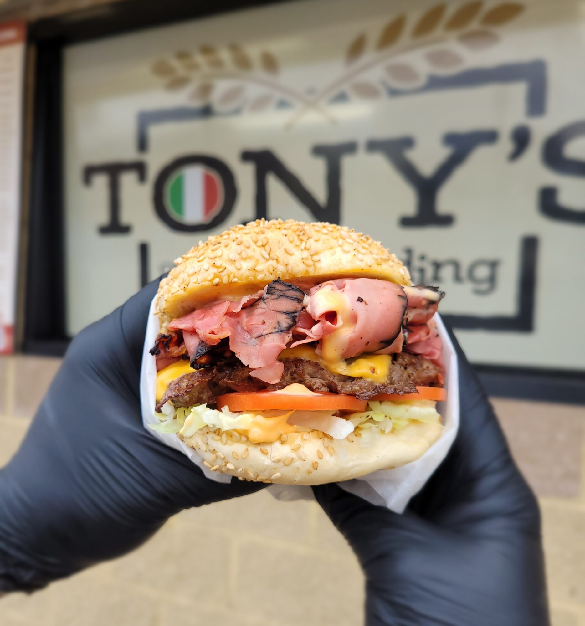 Person wearing black gloves holding a sandwich with meat, cheese, and vegetables outside a restaurant with a sign in the background.