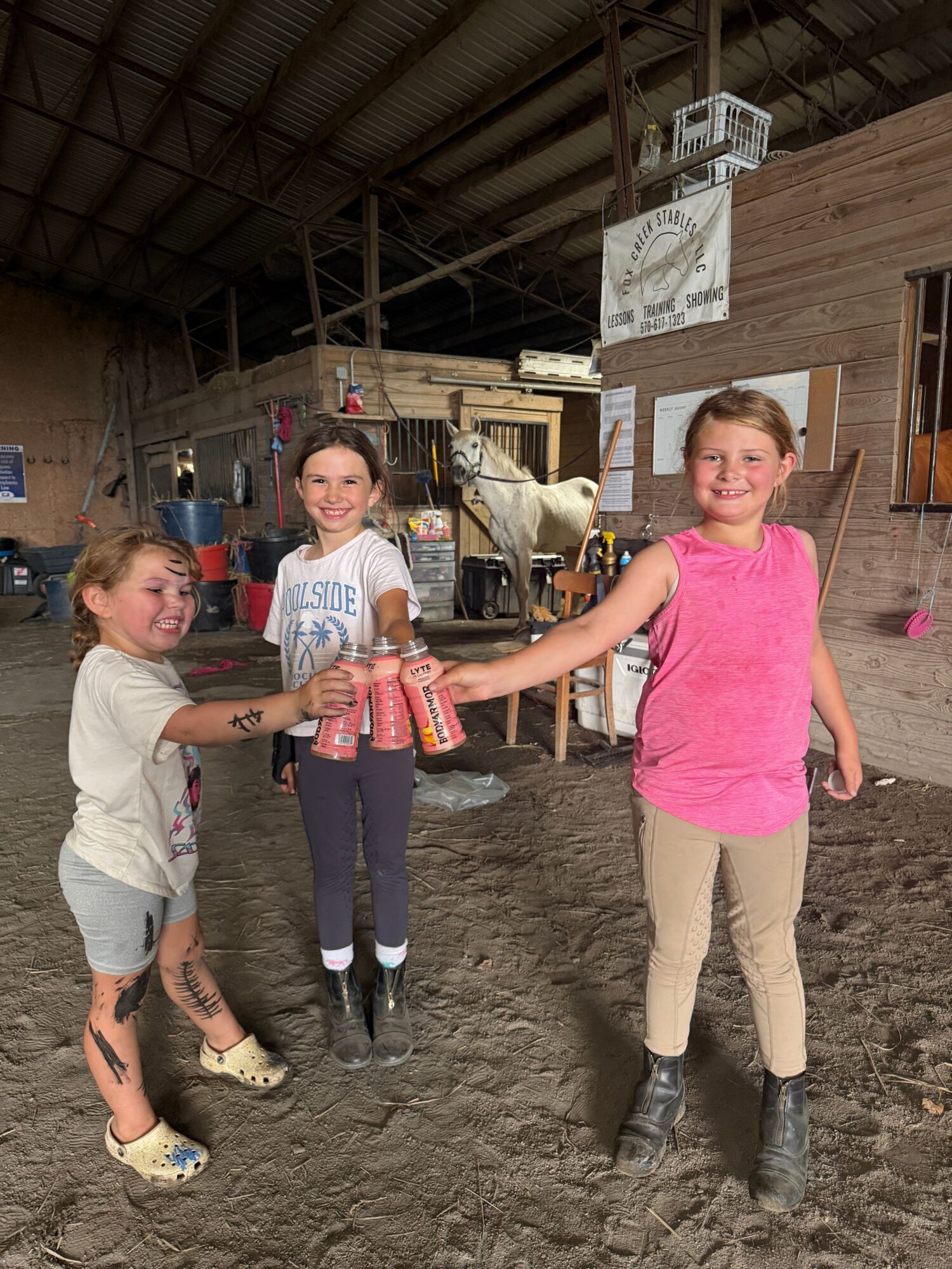 Two women and a girl holding a stack of cans in a barn or warehouse with a dirt floor.