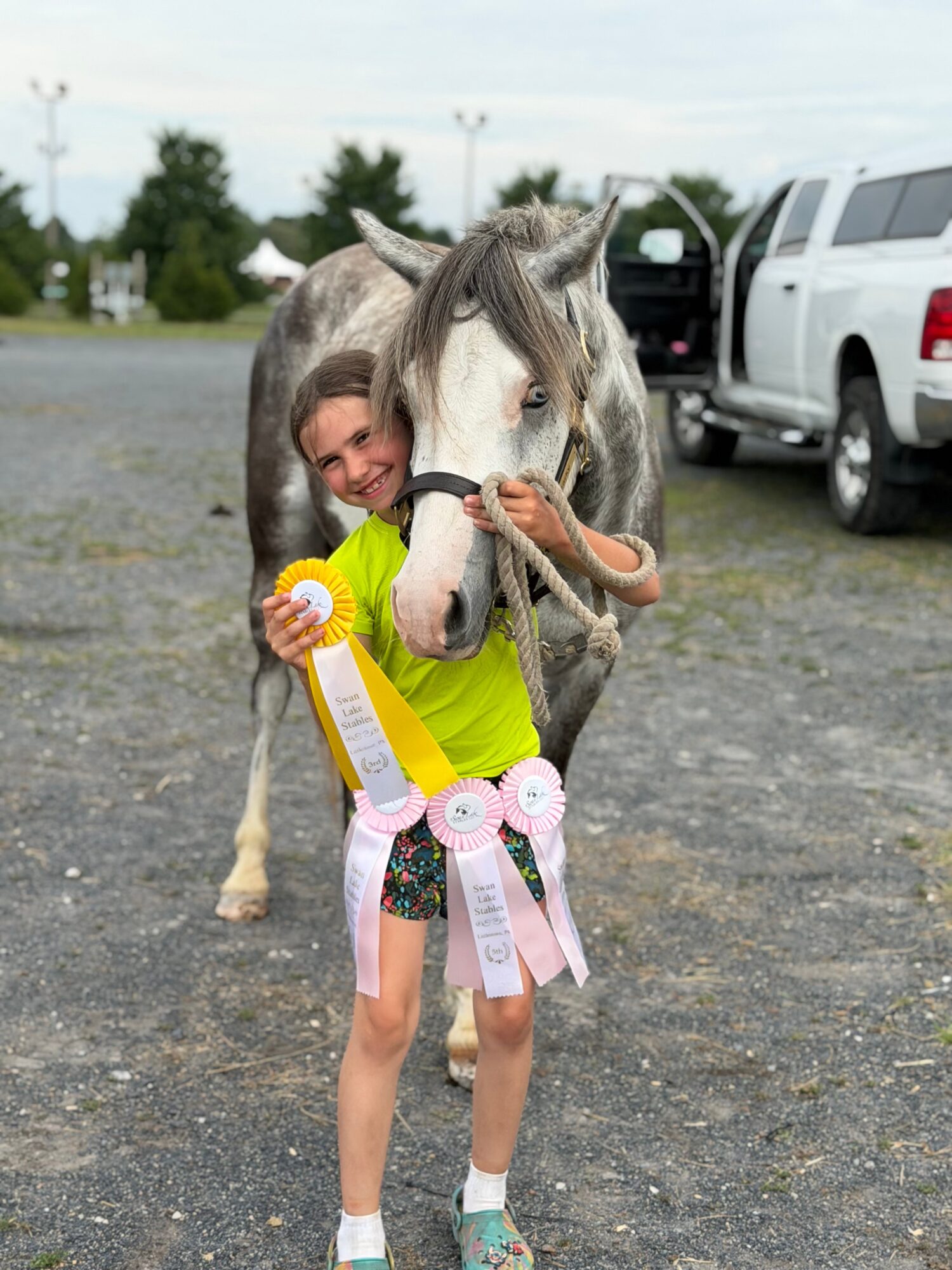 Child hugging a white horse outdoors, smiling, with ribbons attached to her clothing, vehicles in the background.