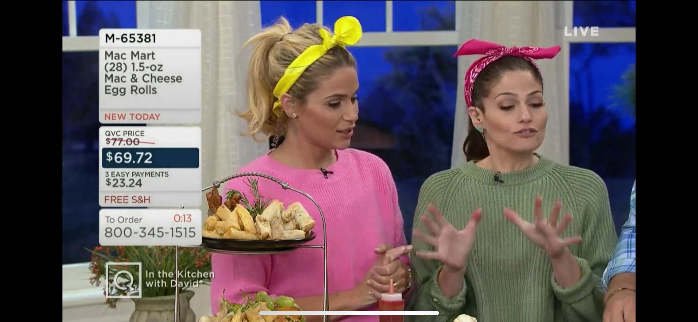 Two women in colorful sweaters and headbands talk on a TV show set with food items and product info displayed.