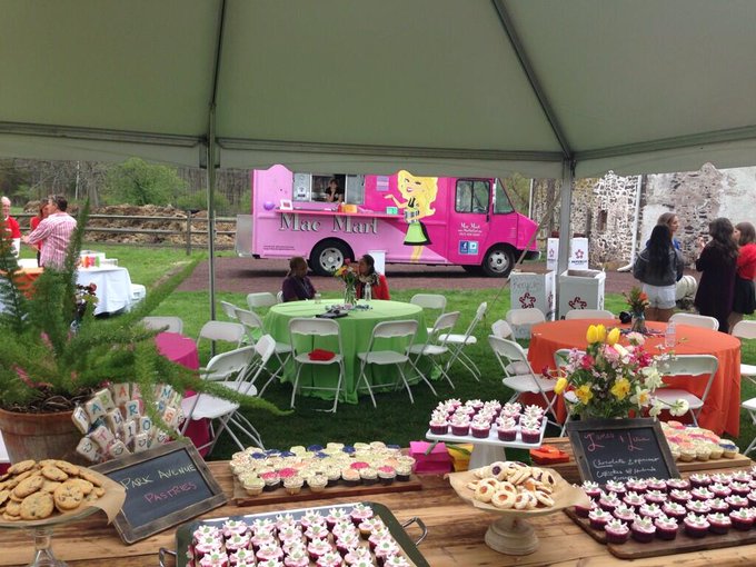 Food and dessert tables under a tent with people in the background, pink food truck, and outdoor setting.